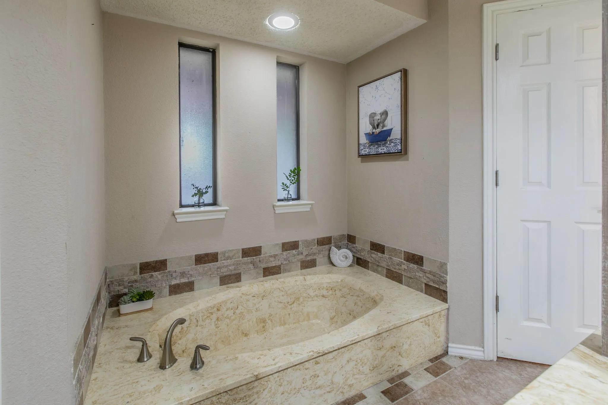 Built-in tub with stone-finish surround, accompanied by dual vertical accent windows, recessed ceiling lighting, and a neutral wall palette