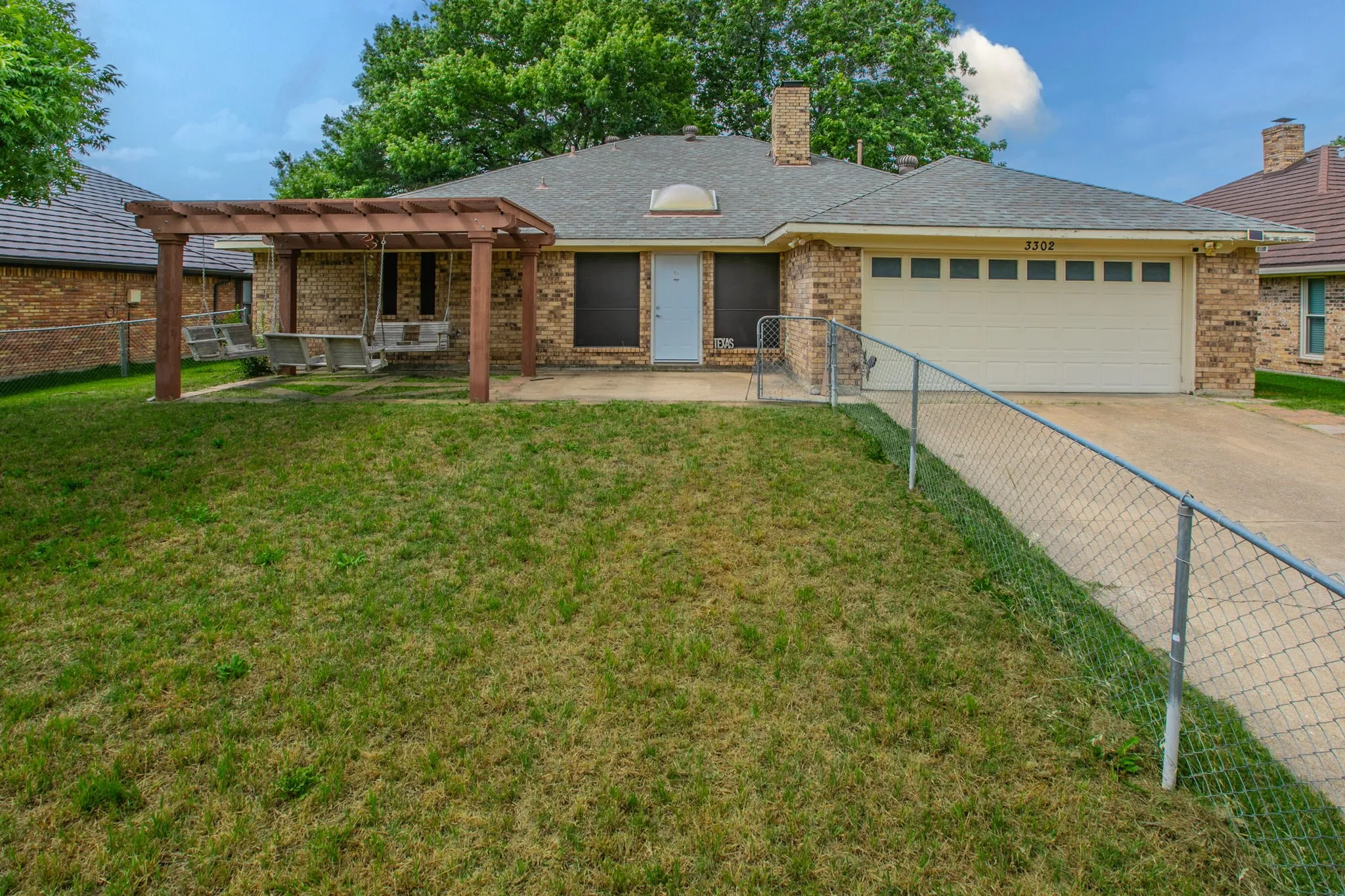 Brick exterior with a prominent front yard, an attached garage, a driveway, a pergola structure, and mature trees