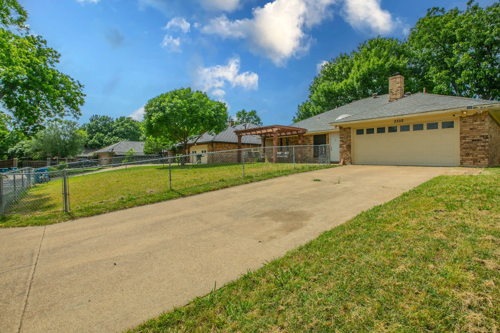 Brick exterior with an attached two-car garage, concrete driveway, chain-link fencing, mature trees, and a wooden pergola structure