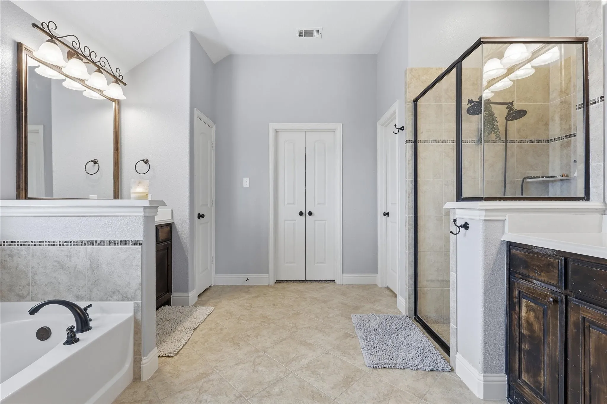 Bathroom featuring a glass-enclosed shower with dual shower heads, a separate soaking tub with dark bronze fixtures, and two distinct vanity areas with wood cabinetry and light countertops