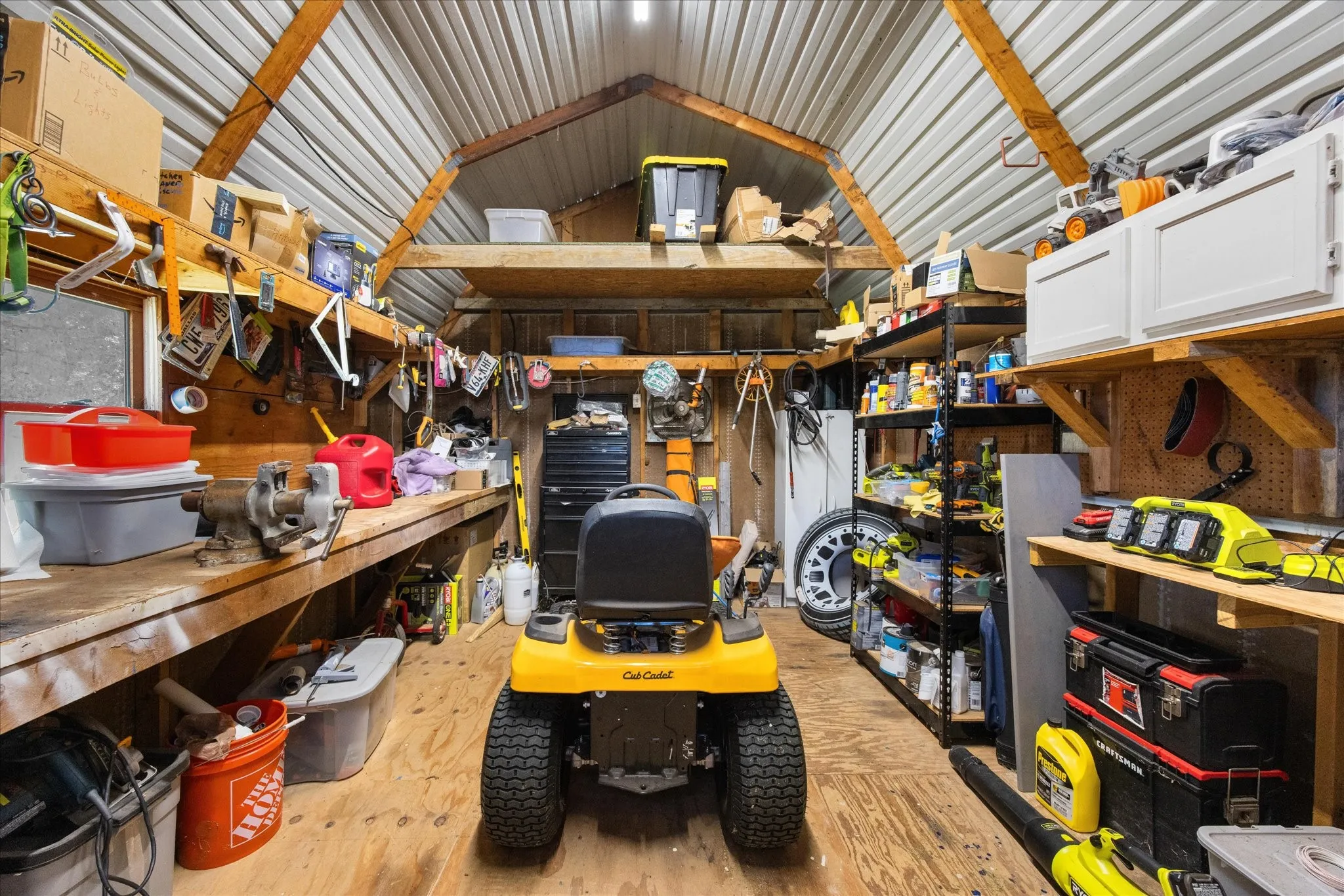 Detached outbuilding with exposed wood framing, corrugated metal ceiling, extensive workbench, built-in shelving, and white cabinetry
