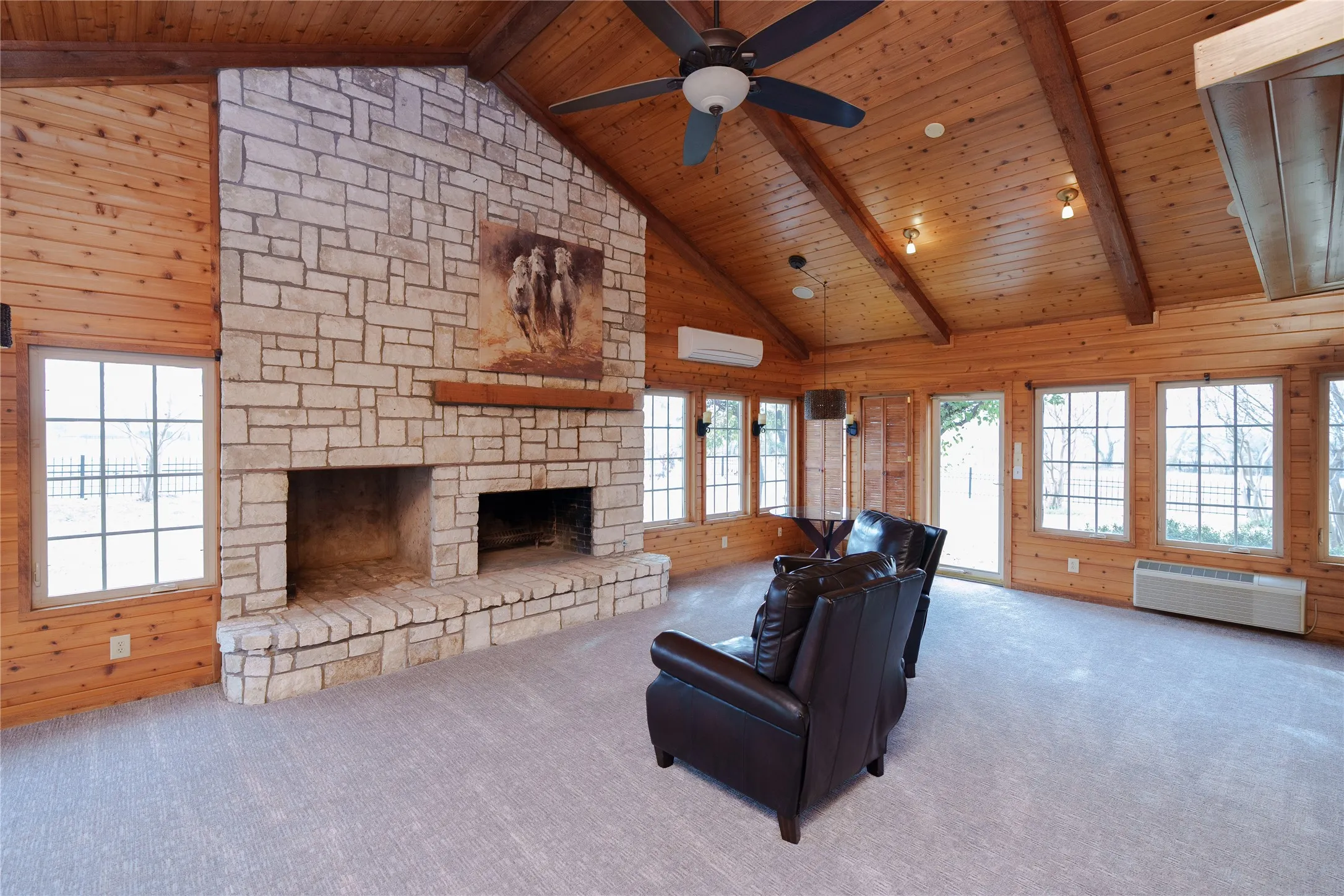Spacious room featuring a prominent stone fireplace with a raised hearth, vaulted wood-paneled ceiling with exposed beams, multiple grid-pattern windows, and a ceiling fan with integrated lighting