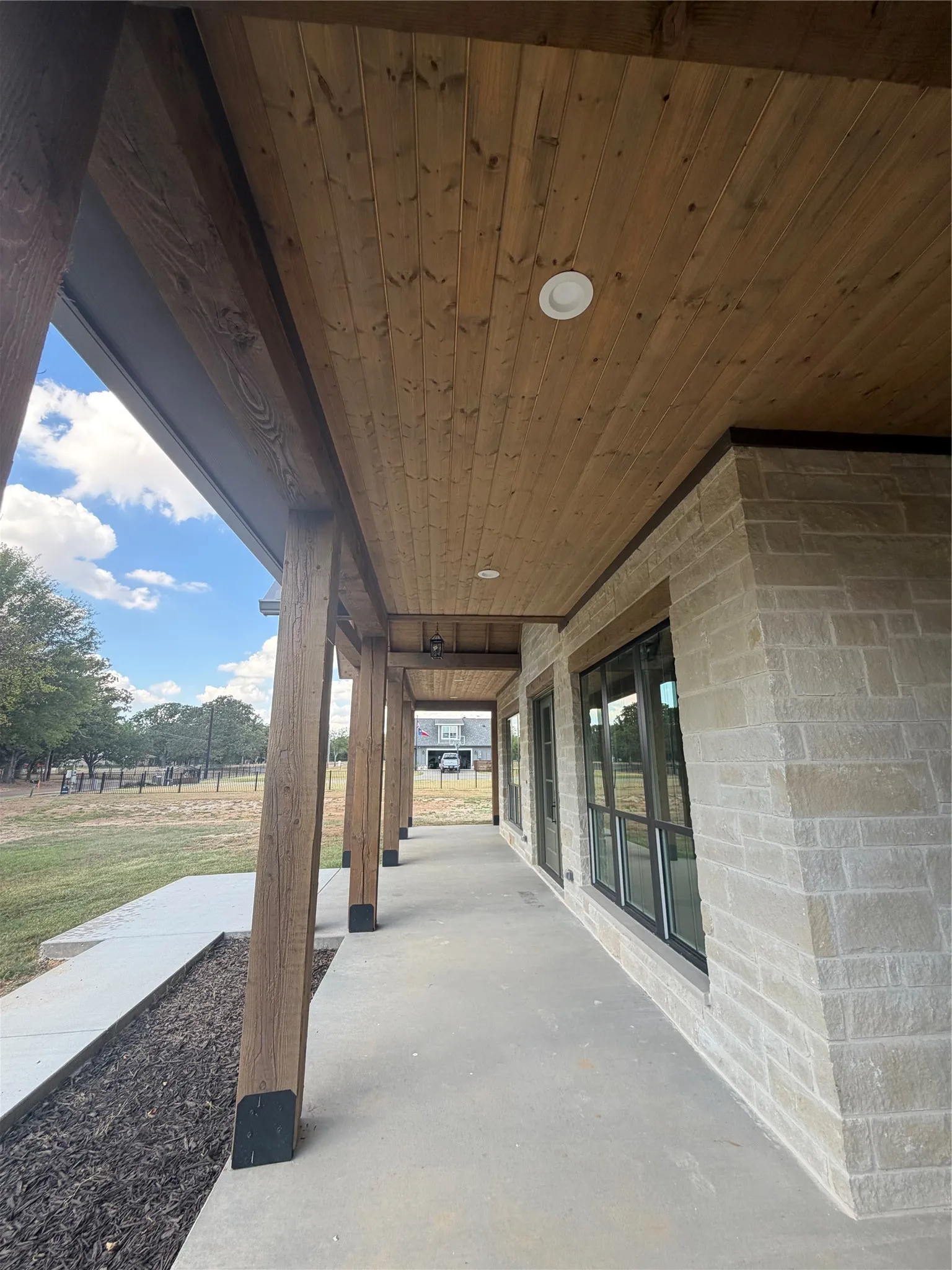 Covered exterior walkway featuring a wood-panel ceiling with recessed lighting, stone veneer siding, and multiple large windows with dark framing