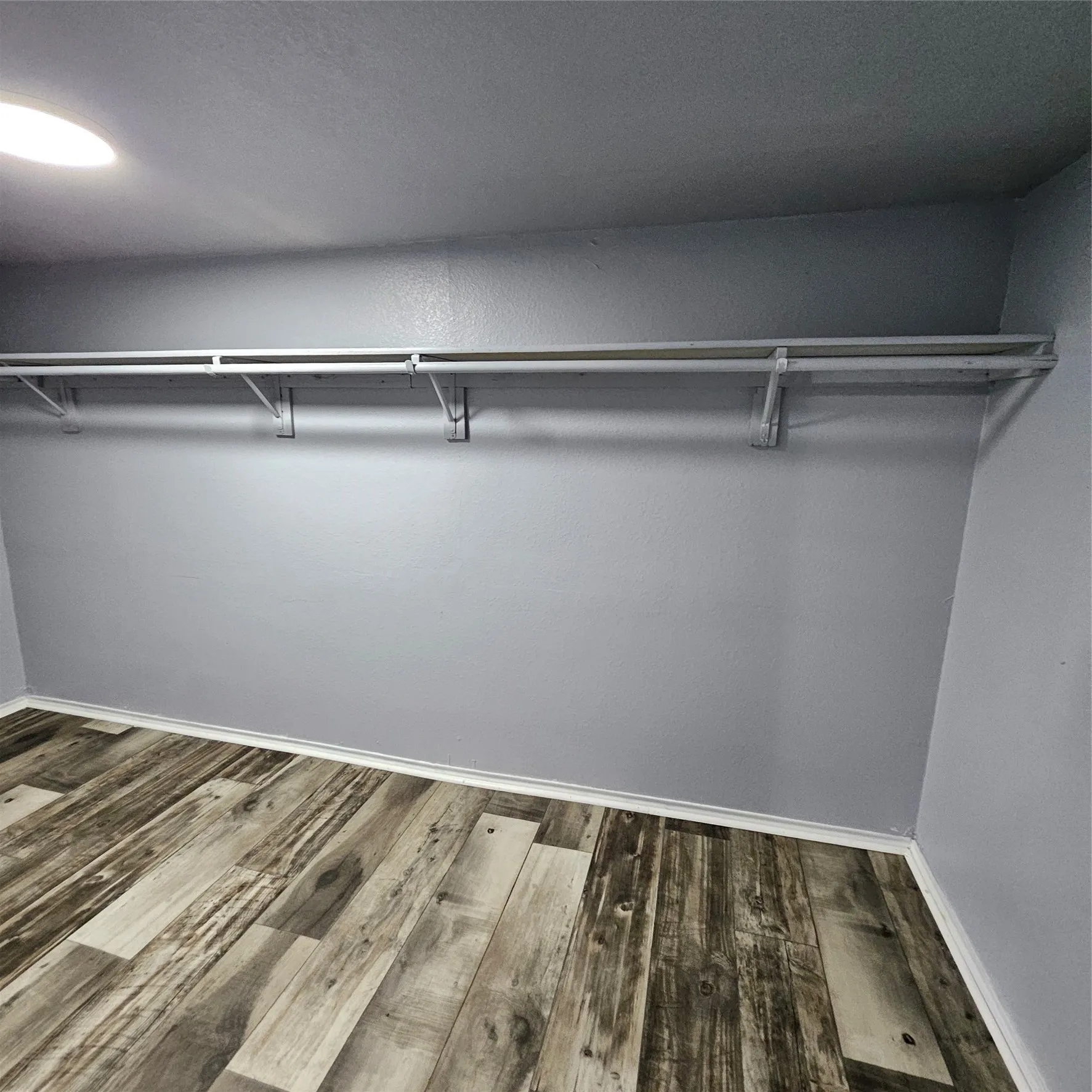 Built-in closet organization system featuring a single hanging rod and upper shelf, set against light gray walls with white baseboards, and wood-finish flooring