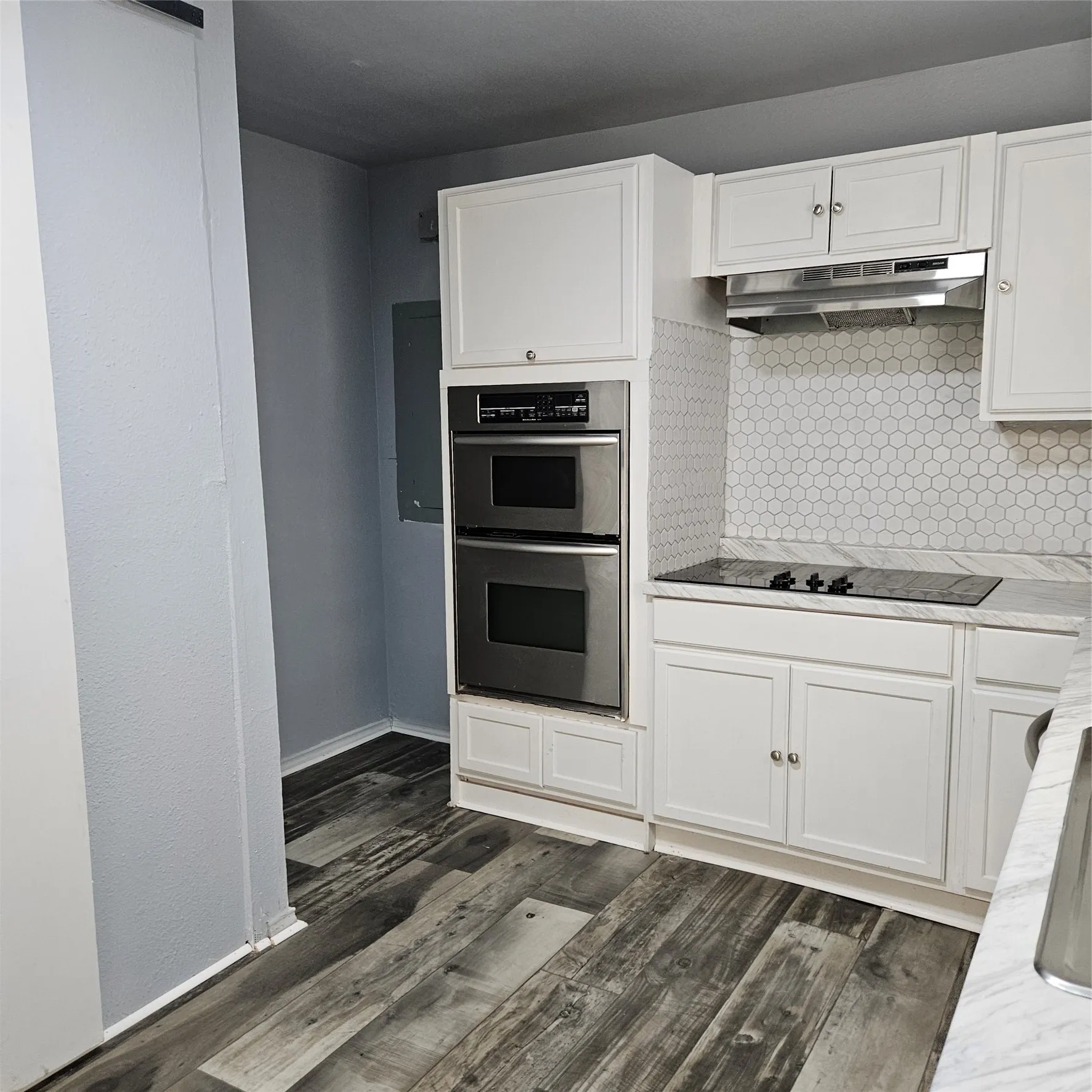 Kitchen featuring white shaker-style cabinetry, a white hexagonal tile backsplash, stainless steel double wall ovens, a black glass cooktop, and wood-finish flooring