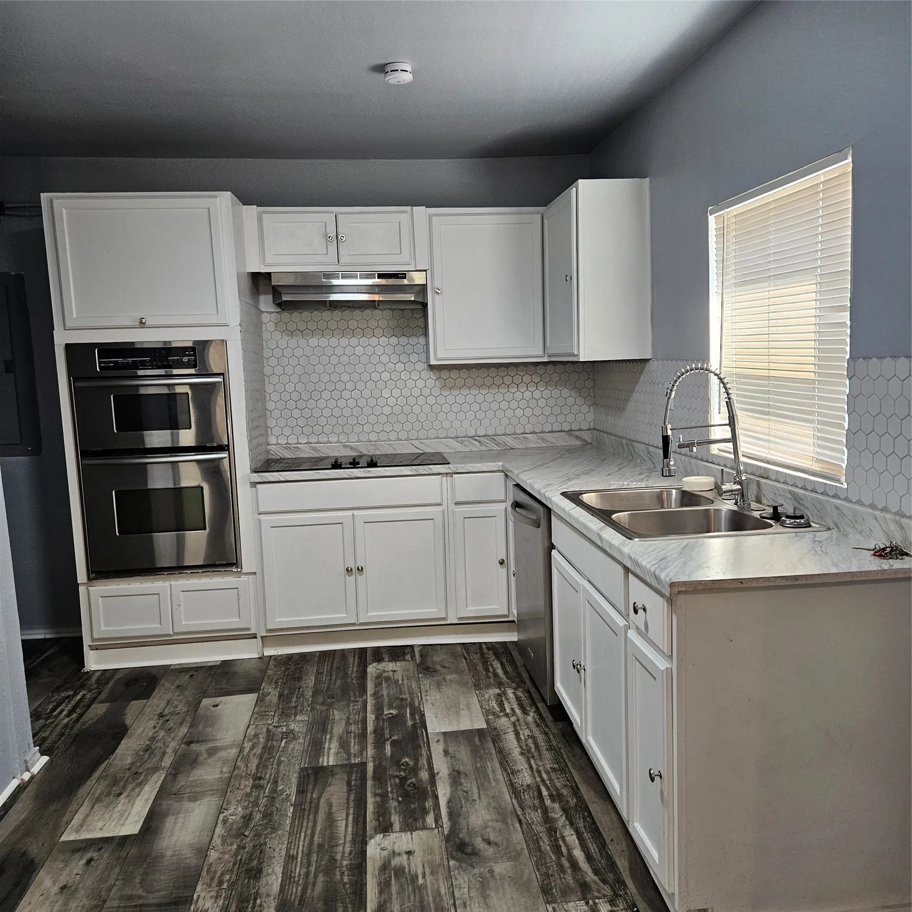 Modern kitchen featuring white shaker cabinetry, an integrated double oven, a stainless steel double sink with a high-arc faucet, and hexagon tile backsplash