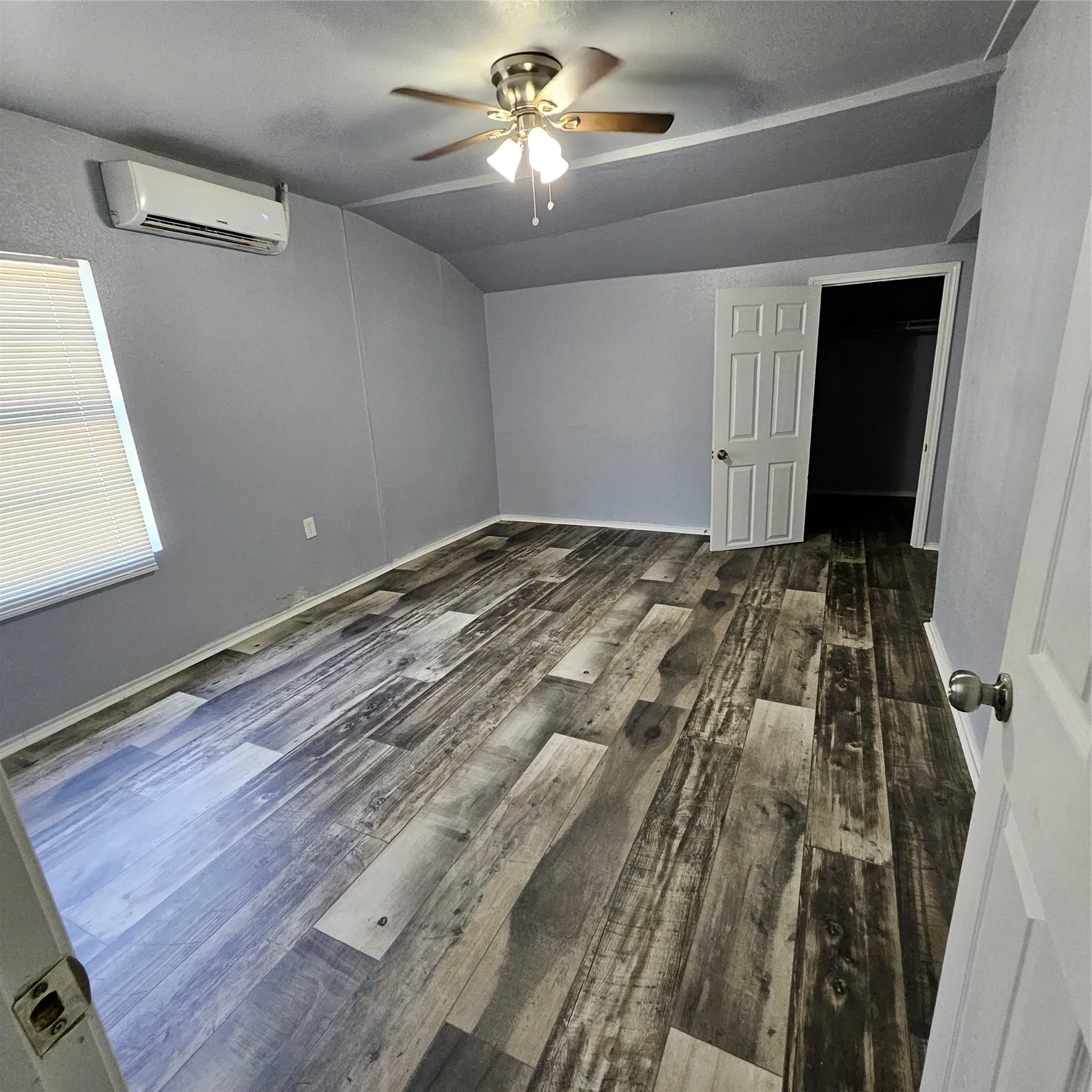 Spacious room featuring wood-finish flooring, a ceiling fan with integrated lighting, a wall-mounted climate control unit, a window with blinds, and a paneled door