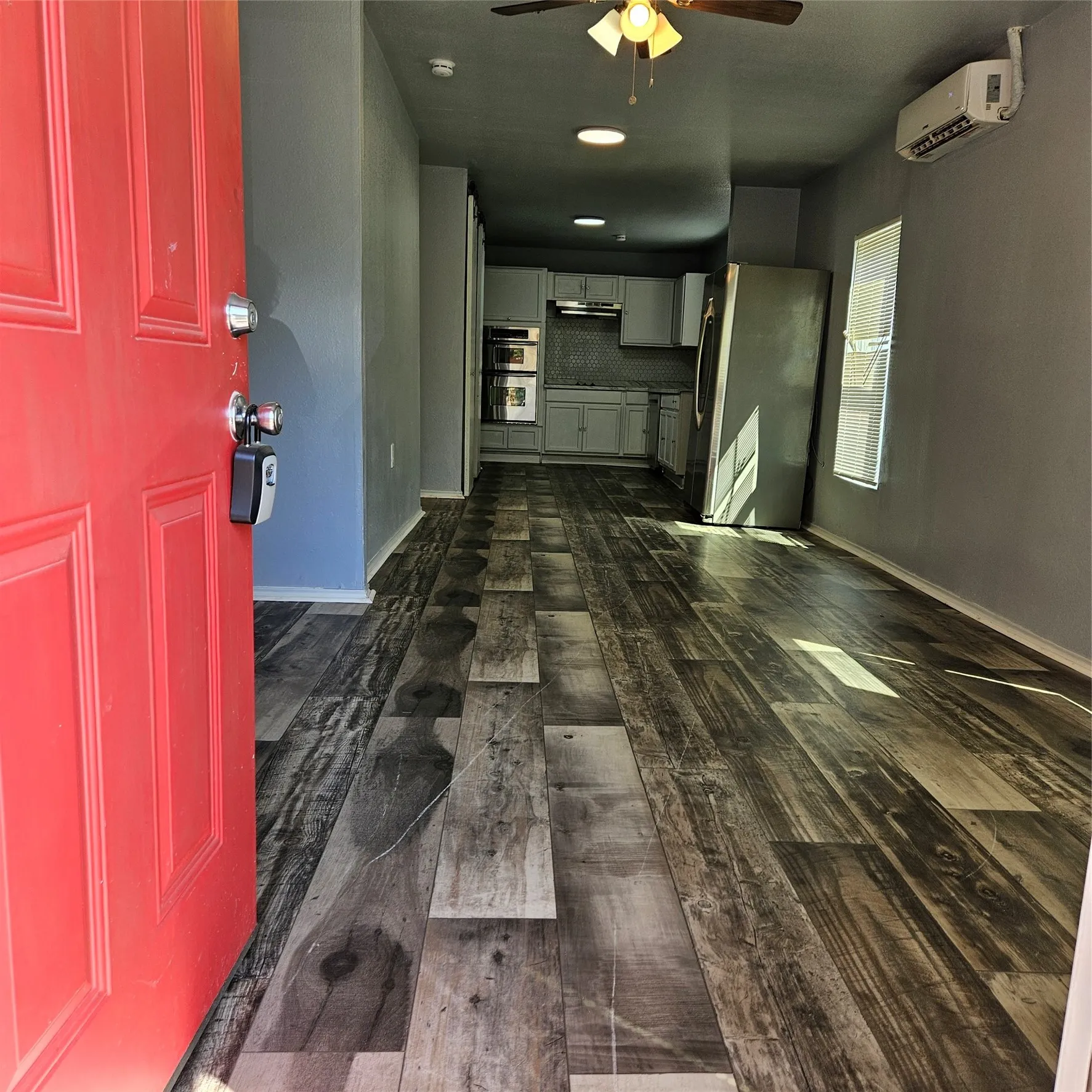 Vibrant red entry door opening to an interior featuring wood-finish flooring, a ceiling fan with integrated lighting, and a modern kitchen with light cabinetry and a tiled backsplash