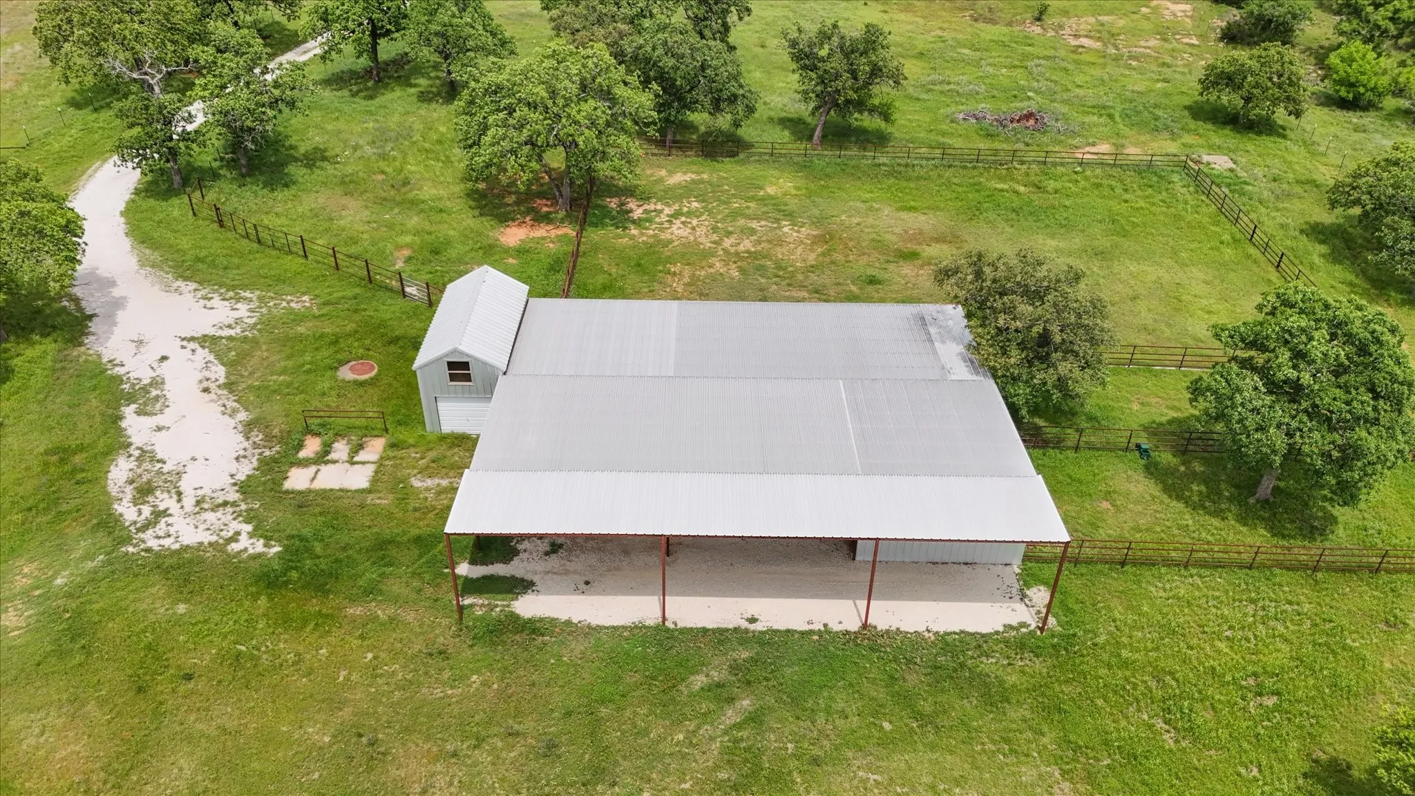 Large metal barn with an attached shed, surrounded by expansive green grounds with mature trees and perimeter fencing