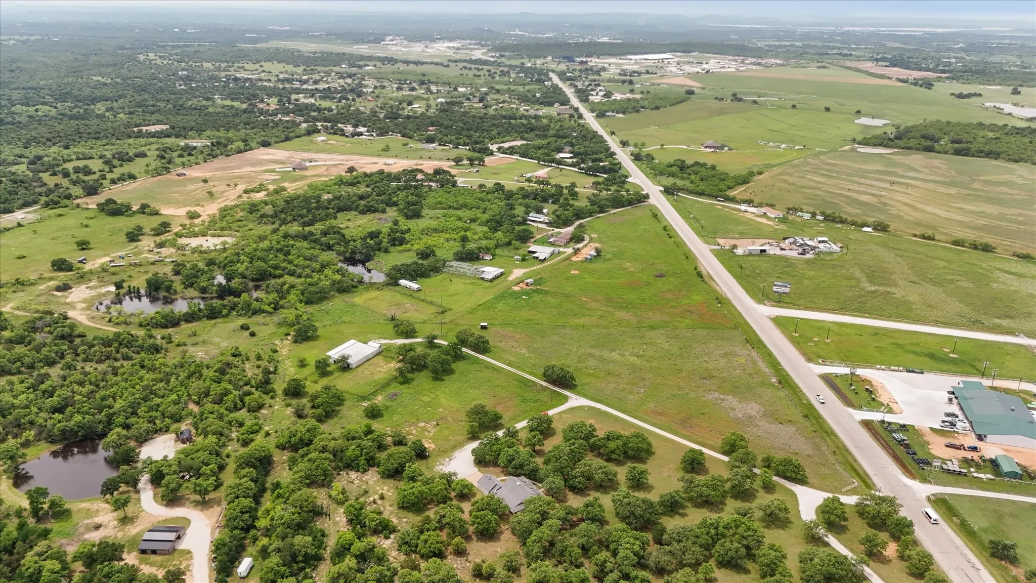 Expansive aerial landscape featuring a prominent paved road dissecting lush green fields and wooded areas