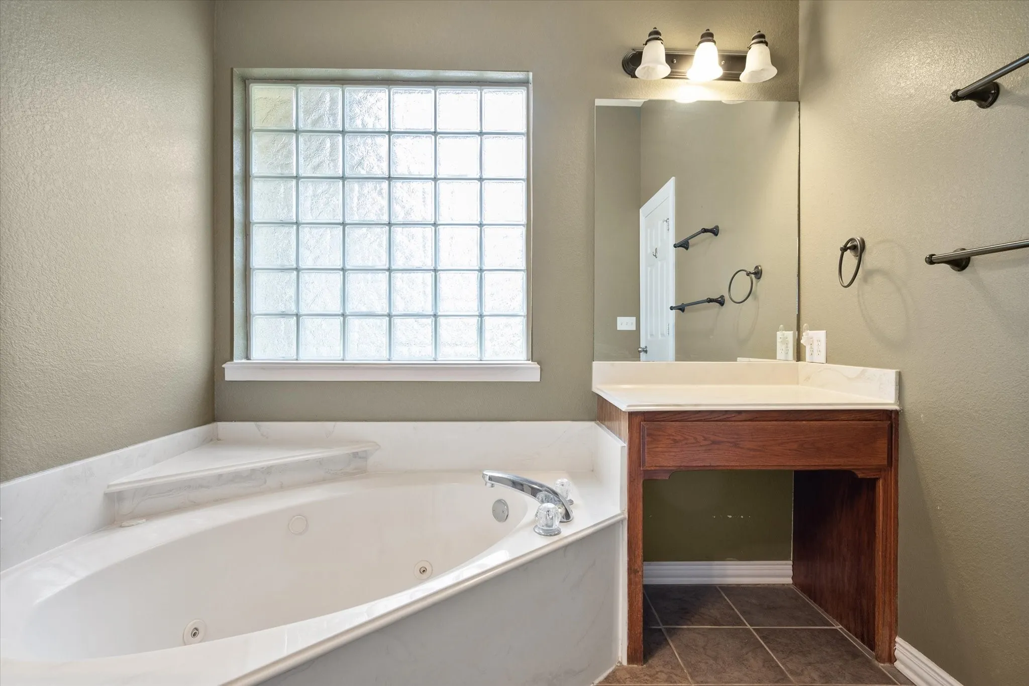 Corner soaking tub with a tiled surround, glass block window, and an adjacent vanity with a light-colored countertop and a wood-finish base