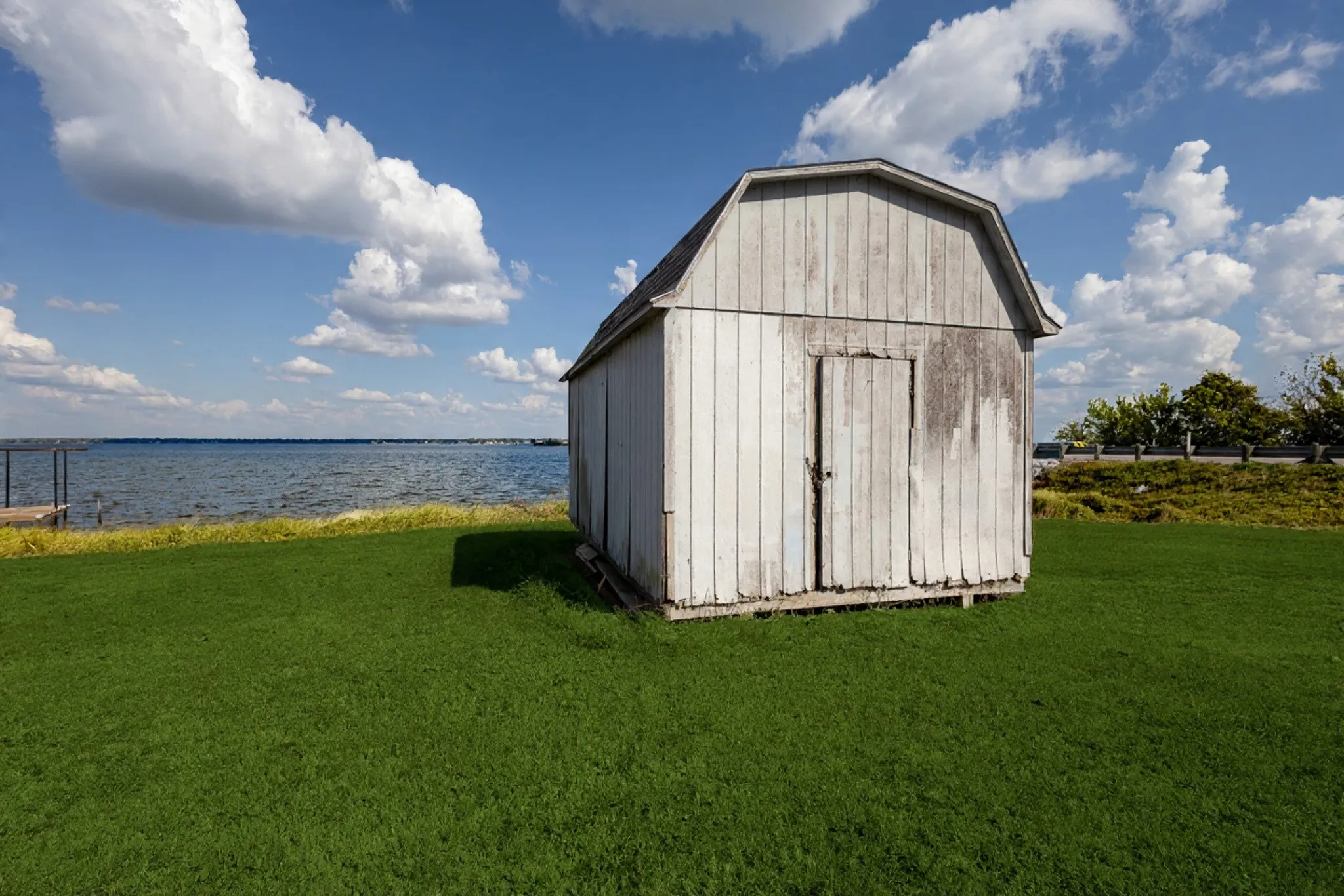 Waterfront property featuring a gambrel-roofed shed, expansive green lawn, and a dock structure