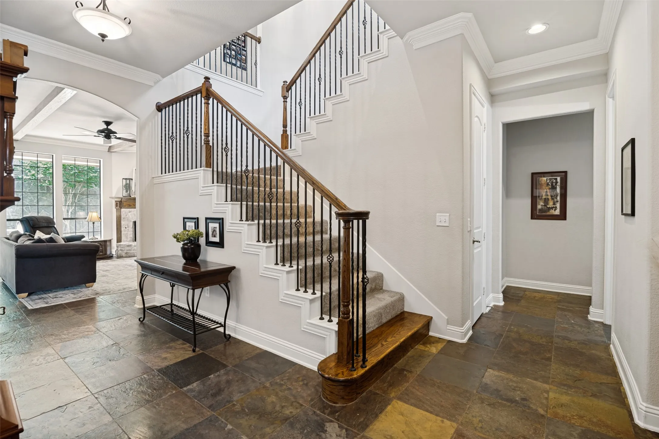 Grand entryway featuring variegated slate flooring, an elegant staircase with wrought iron balusters and wood handrails, a coffered ceiling with crown molding, and a spacious archway opening to a living area with large windows
