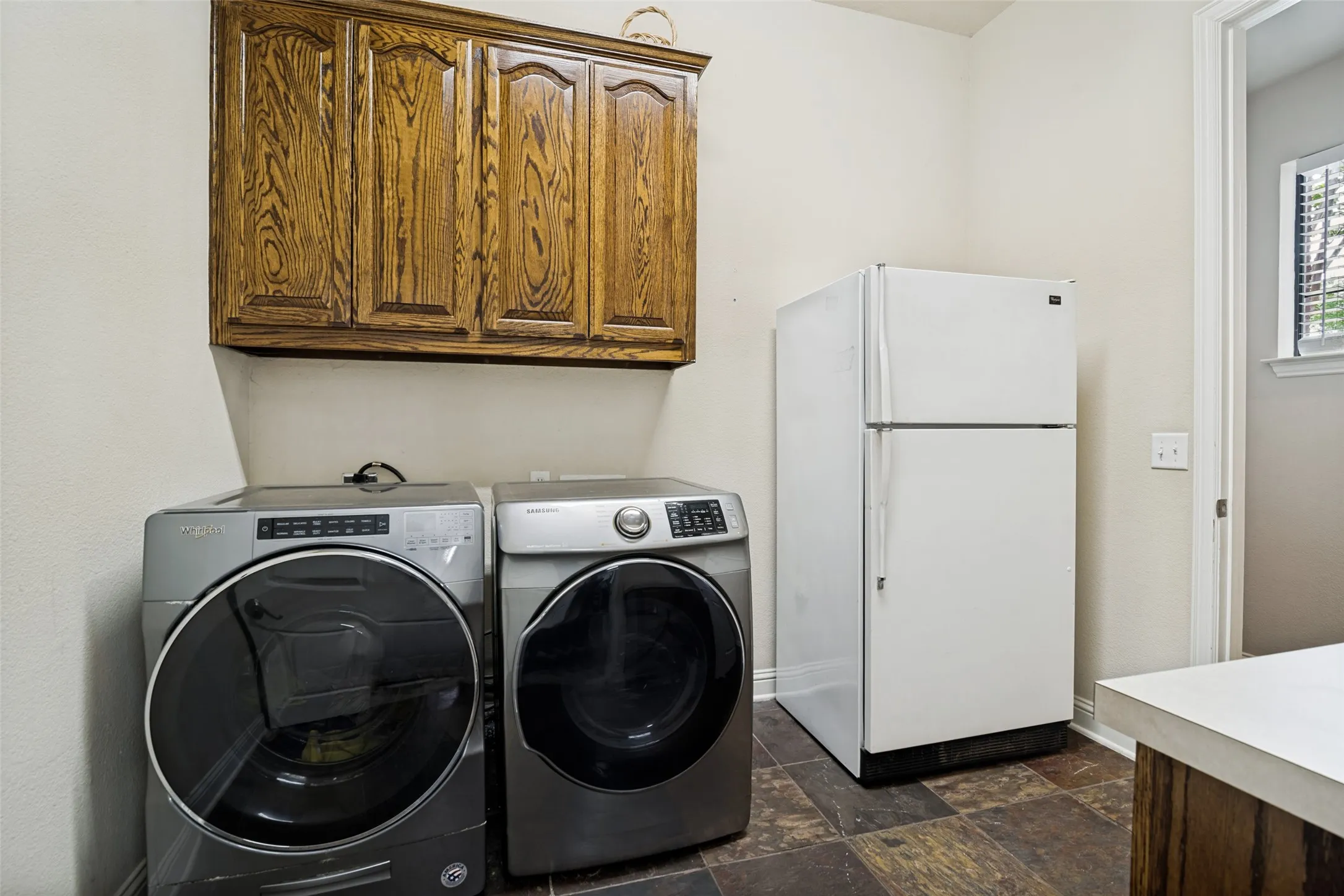 Roomy laundry area featuring upper wood cabinetry, a white refrigerator, and variegated brown floor tiling