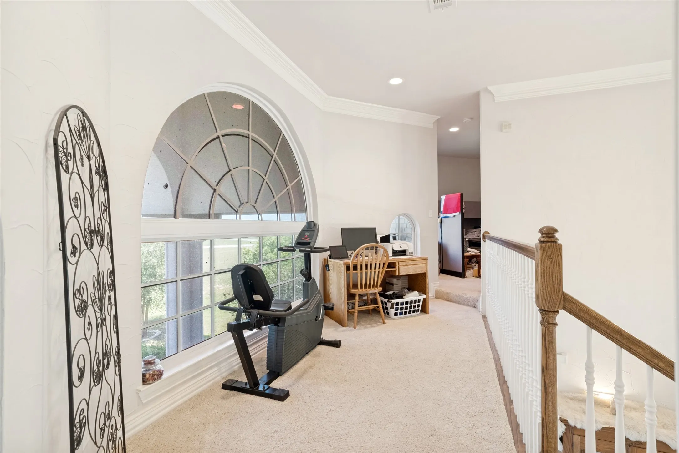 Carpeted loft area featuring a large arched window with grid detailing, crown molding, and a wood banister with white spindles
