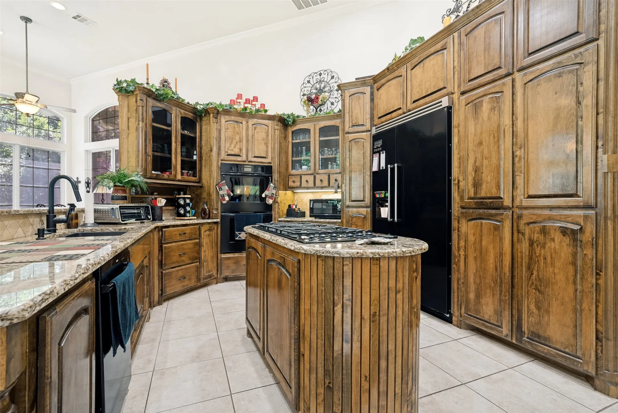 Curved kitchen with extensive wood cabinetry, stone countertops, a large center island with cooktop, integrated double ovens, and a built-in black refrigerator
