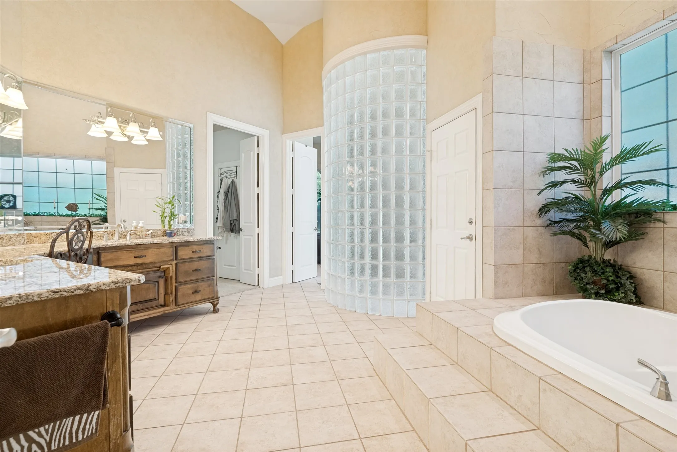 Spacious bathroom featuring a built-in soaking tub, a curved glass block shower enclosure, a dual vanity with a granite countertop, and tiled flooring