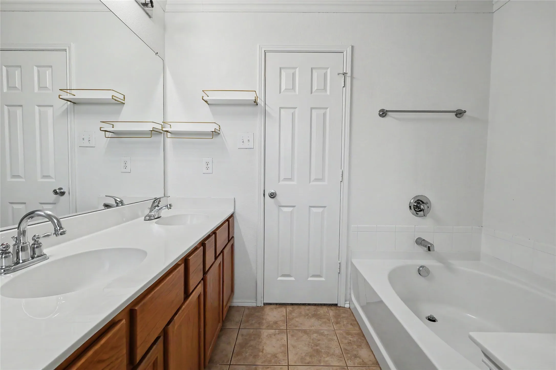 Bathroom featuring a dual-sink vanity with wood-finish cabinetry, a large integrated mirror, a full-size bathtub with white tile surround, and neutral floor tiling