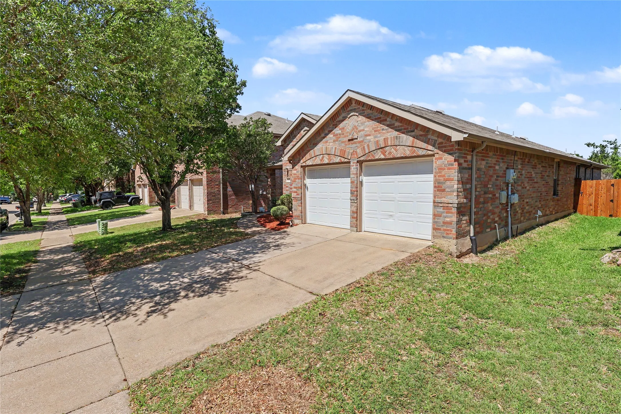Brick exterior featuring a two-car garage with white paneled doors, concrete driveway, front yard with mature trees, and partial wooden privacy fence