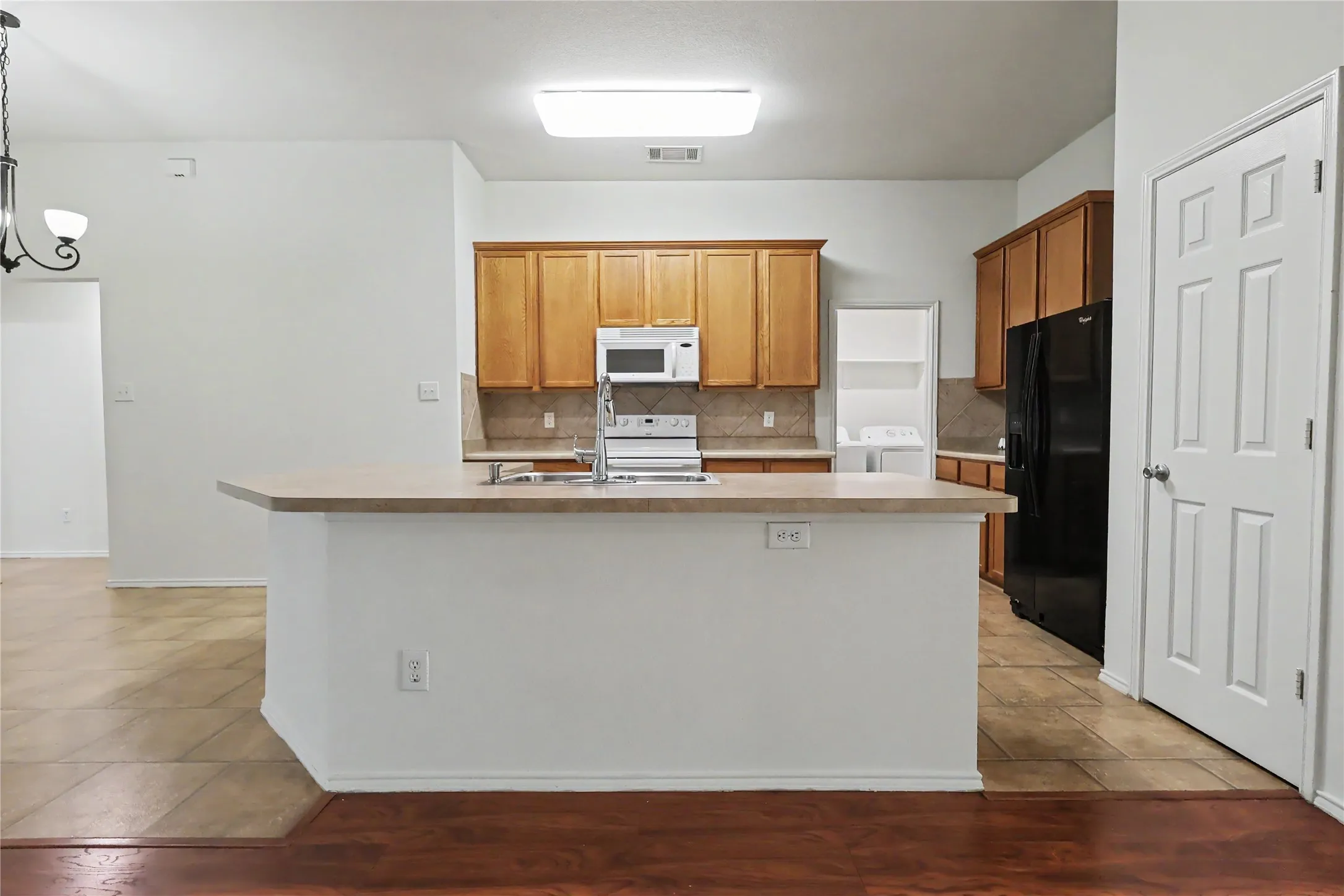 Kitchen island with a sink and chrome faucet, wood cabinetry, tile backsplash, built-in microwave, and wood-finish flooring