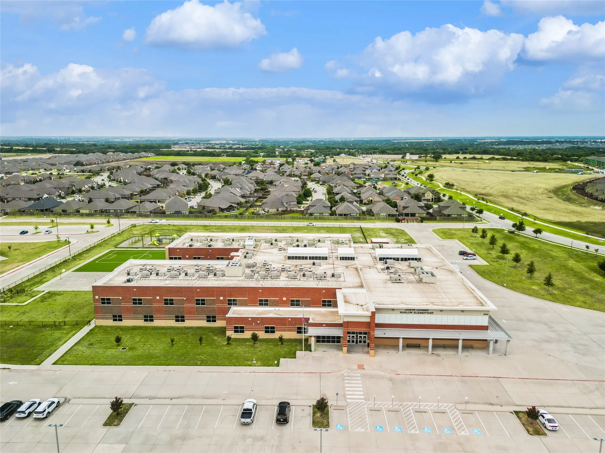 Expansive community showcasing a large institutional building with brick and stone accents, a substantial paved parking area, manicured green spaces, and a surrounding residential neighborhood with numerous single-family homes