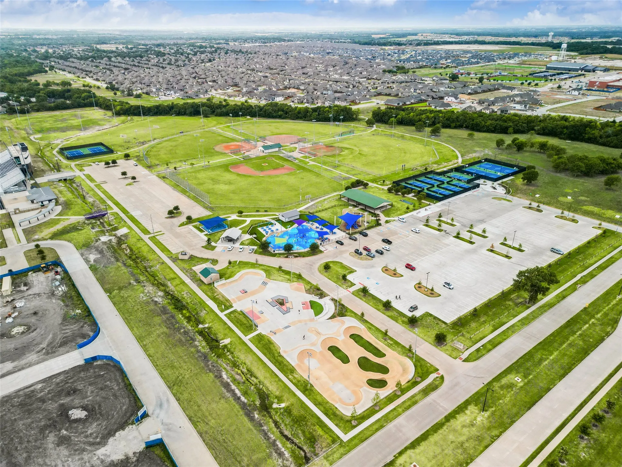 Expansive park setting featuring multiple sports fields, tennis courts, a water play area, a skate park, and extensive paved parking areas
