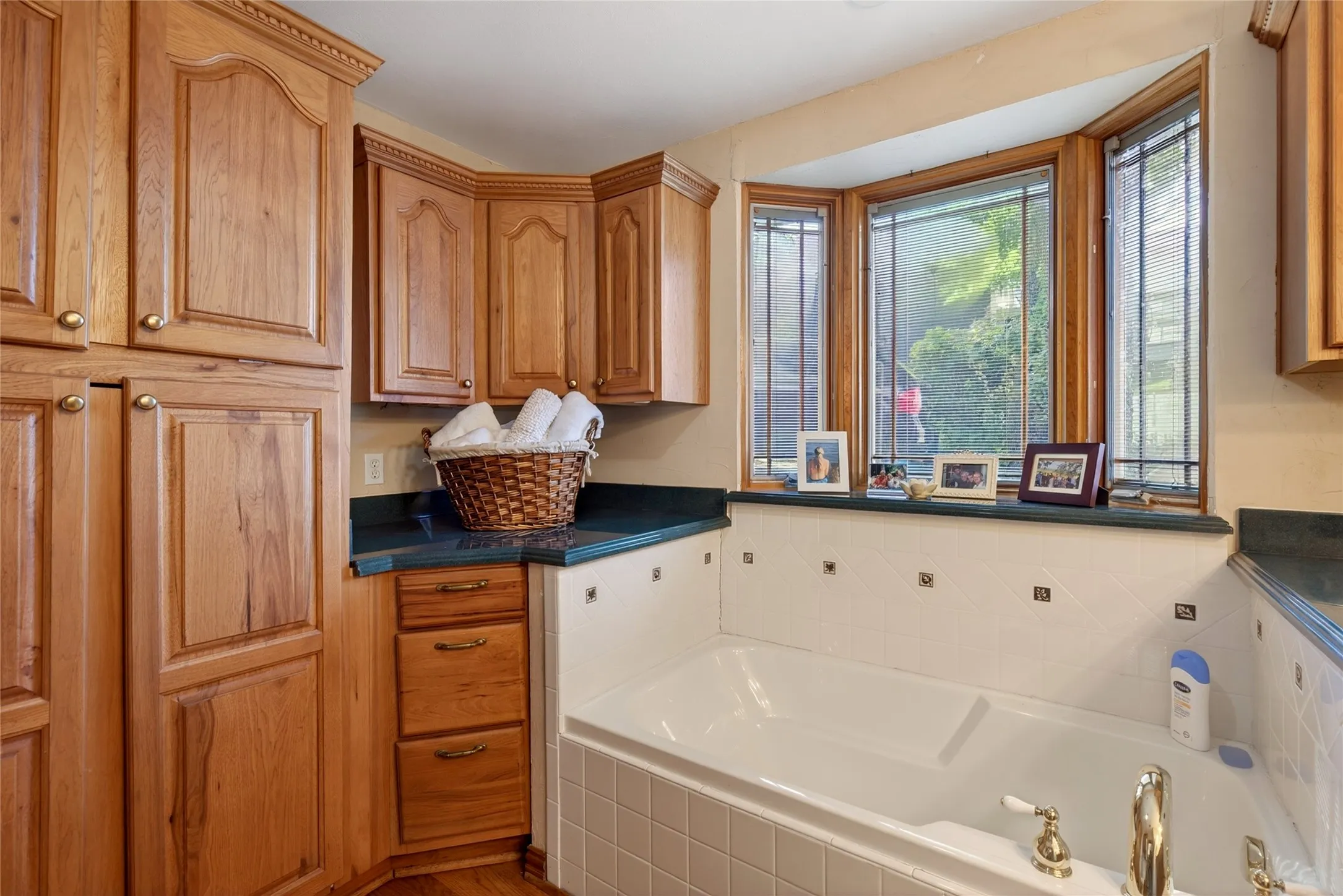 Built-in soaking tub featuring gold-tone fixtures, white tile surround, a bay window with wood-frame construction, with oak cabinetry, and a single sink.