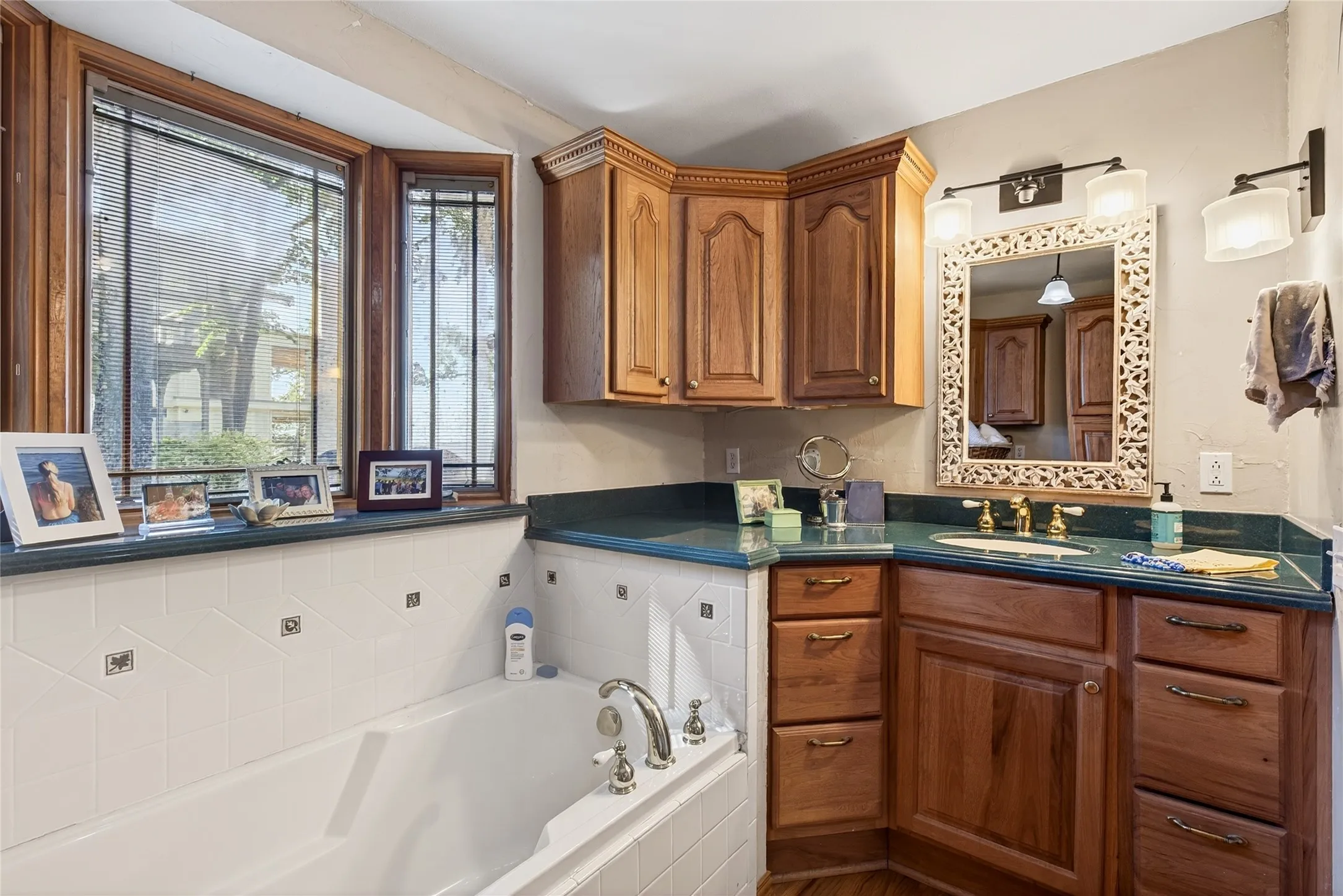 Built-in soaking tub featuring gold-tone fixtures, white tile surround, a bay window with wood-frame construction, with oak cabinetry, and a single sink.