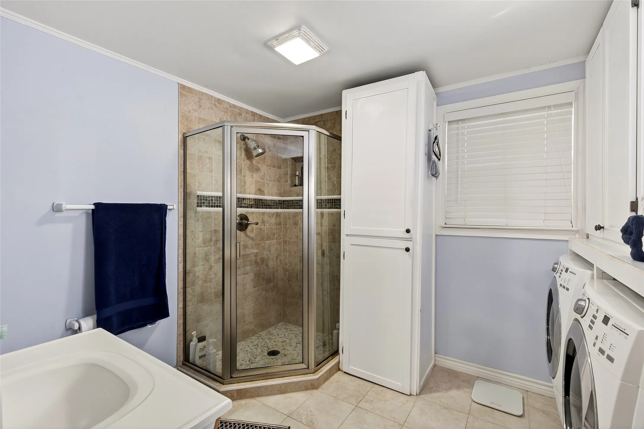 Corner glass-enclosed shower with tiled surround and pebble floor, single sink, cabinets, and washer and dryer.