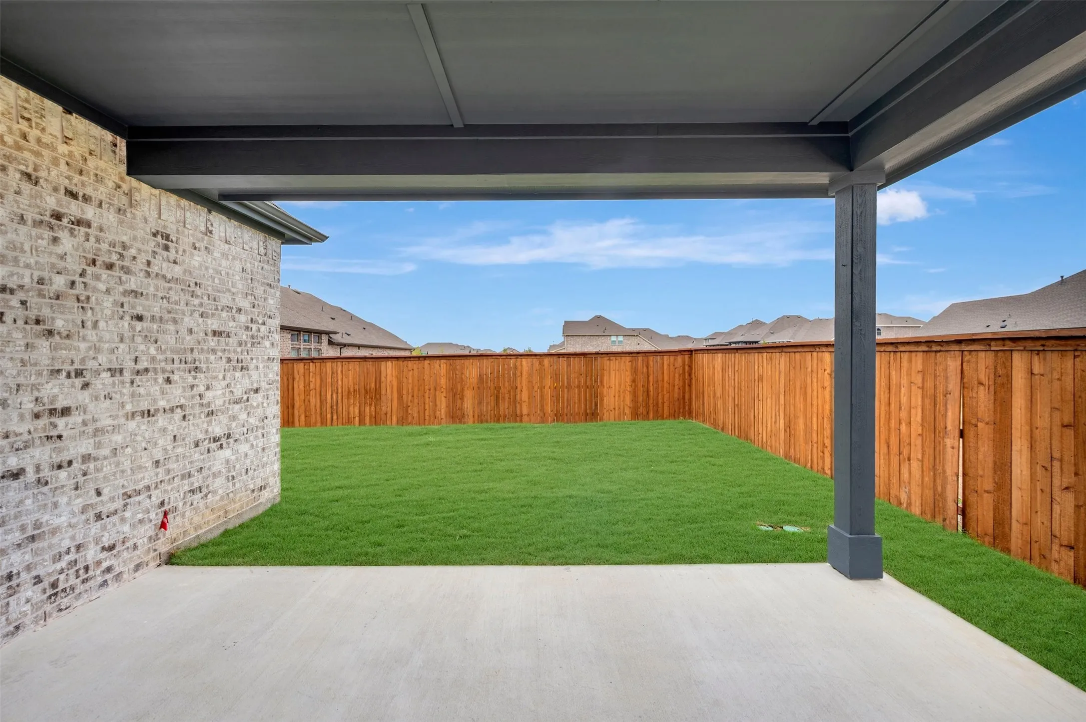Covered patio with concrete flooring, brick exterior, and a fenced backyard with green lawn