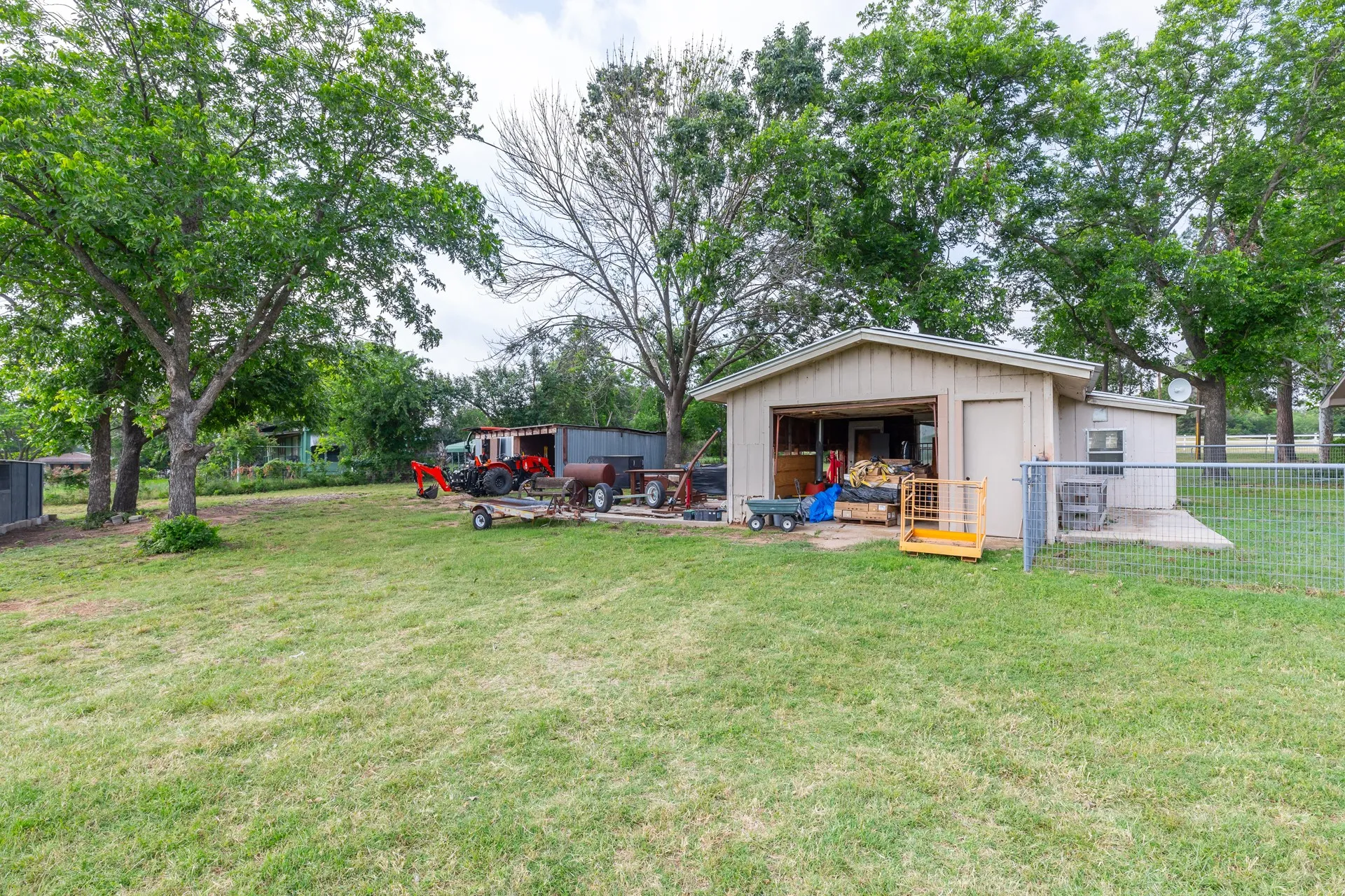 Detached utility building with an open garage door and a side entry door, situated on a grassy lot with mature trees
