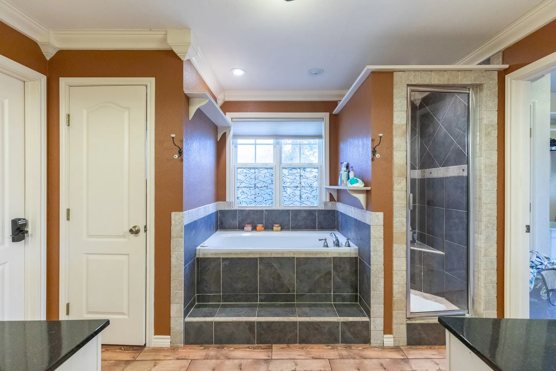 Bathroom featuring a built-in tub with dark tile surround and steps, a tiled walk-in shower with glass enclosure, white trim, and a window with blinds