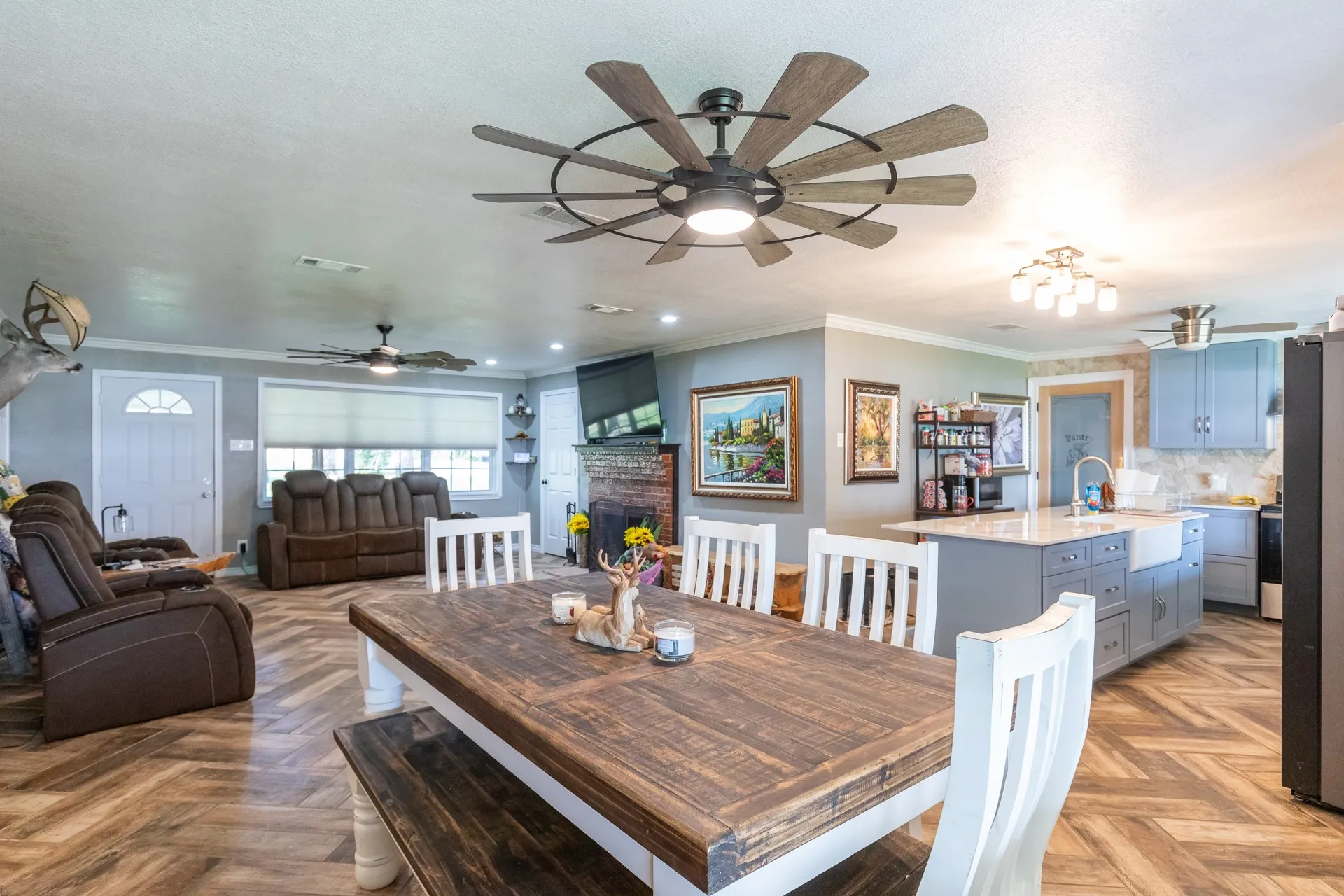 Open-concept living space featuring wood-finish flooring in a herringbone pattern, a dining area with a rustic-style table, a kitchen island with a farmhouse sink, light blue cabinetry, and a brick fireplace