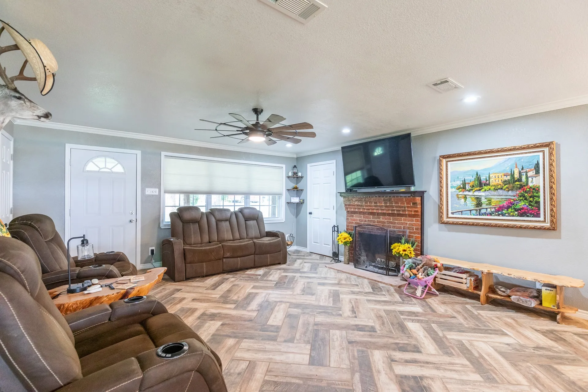 Spacious living area featuring herringbone-pattern wood-finish flooring, a brick fireplace with a hearth, and multiple windows with roller shades