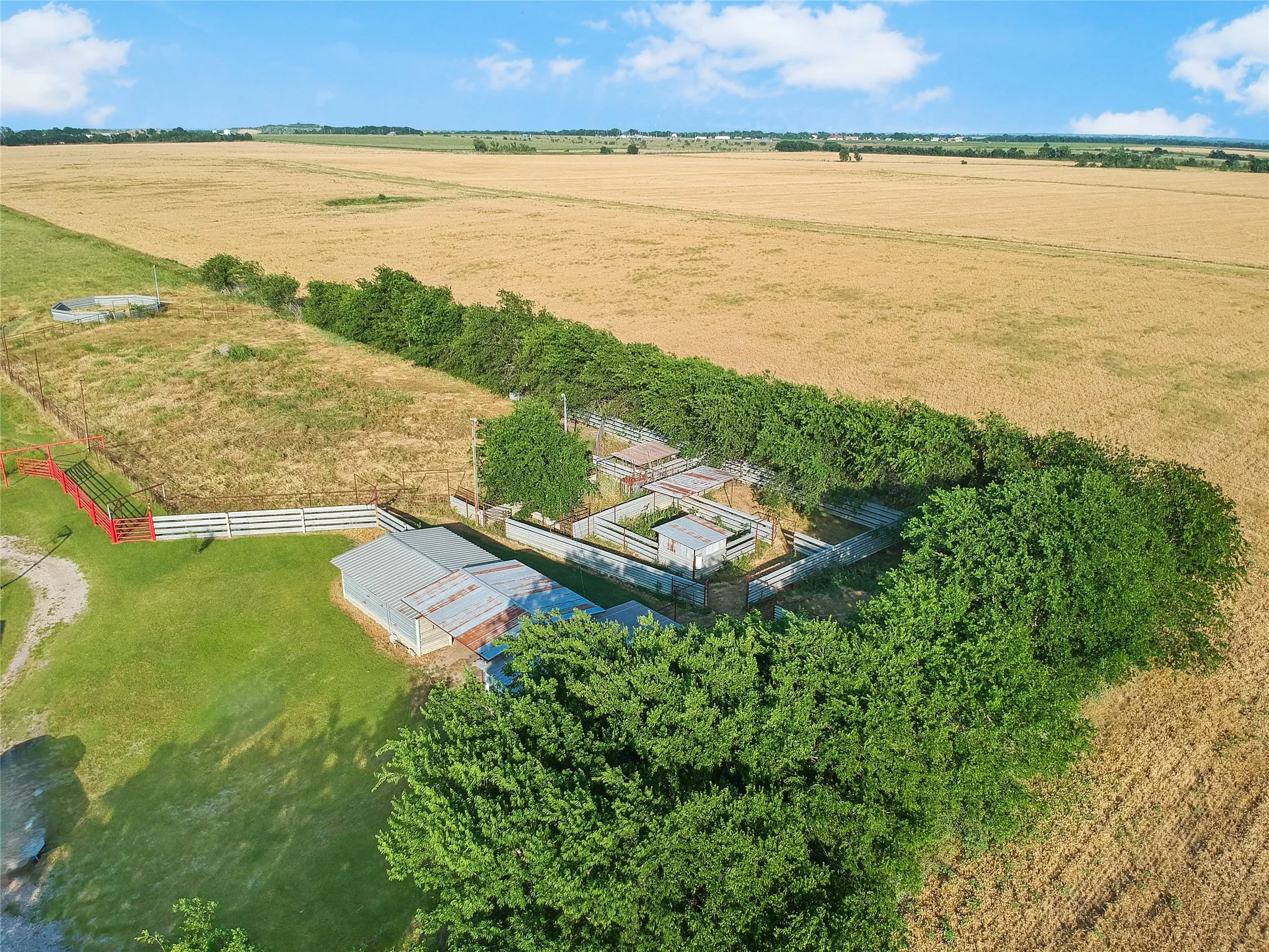 Expansive agricultural property featuring multiple fenced areas, a metal-roofed outbuilding, mature trees, and open fields