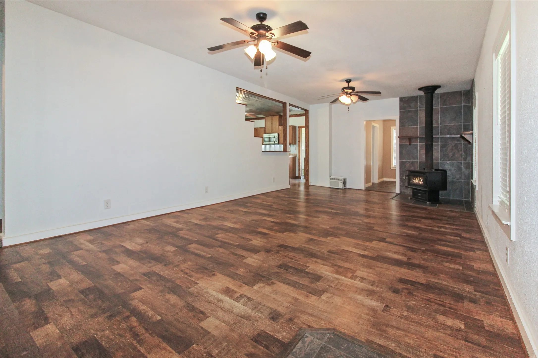 1936  HOUSE Spacious living area featuring wood-finish flooring, a freestanding wood-burning stove with a tiled surround, and multiple ceiling fans with integrated lighting