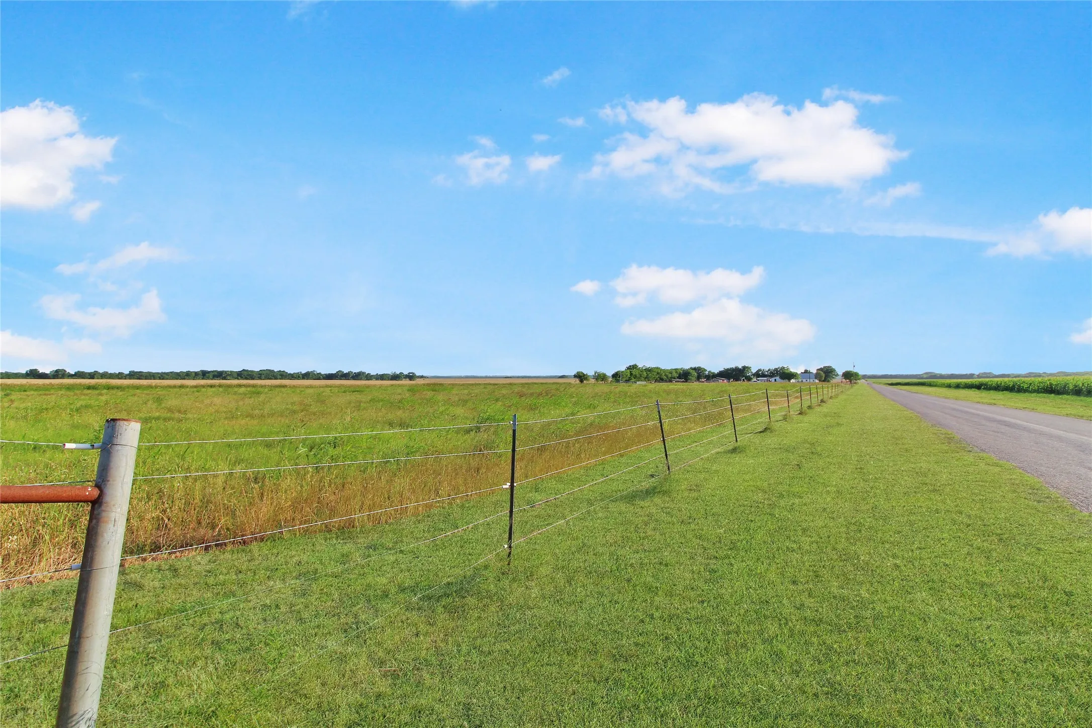 Expansive green fields alongside a paved road, bordered by a wire fence with metal posts
