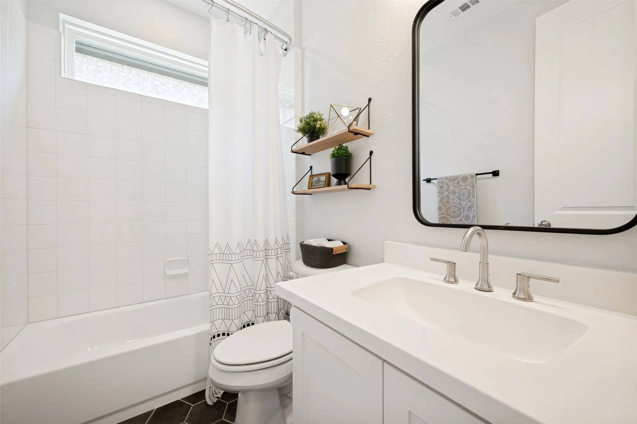 This bright bathroom has a shower/tub combo and modern white aesthetic with trendy dark tile flooring