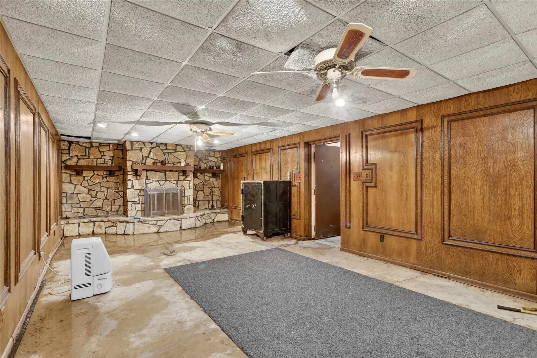 Spacious room down stairs featuring a stone-faced fireplace, wood-paneled walls with decorative trim, and a drop ceiling ceiling fans