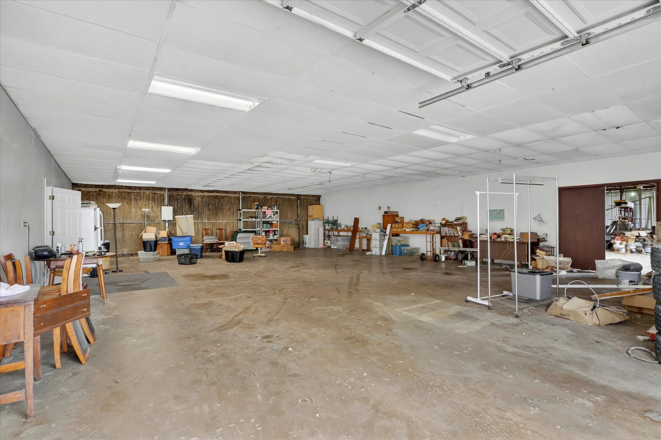 Expansive garage space featuring a concrete floor, drop ceiling with fluorescent lighting, overhead garage door and door leading to larger warehouse space