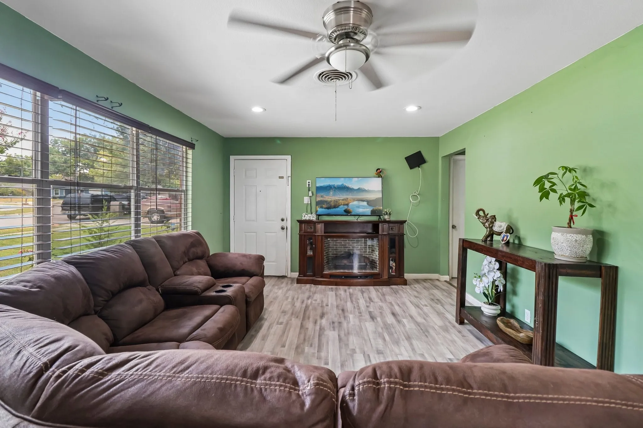 Spacious living room featuring fresh sage green walls, wood-finish flooring throughout, a ceiling fan with integrated lighting, and large multi-pane windows with natural light