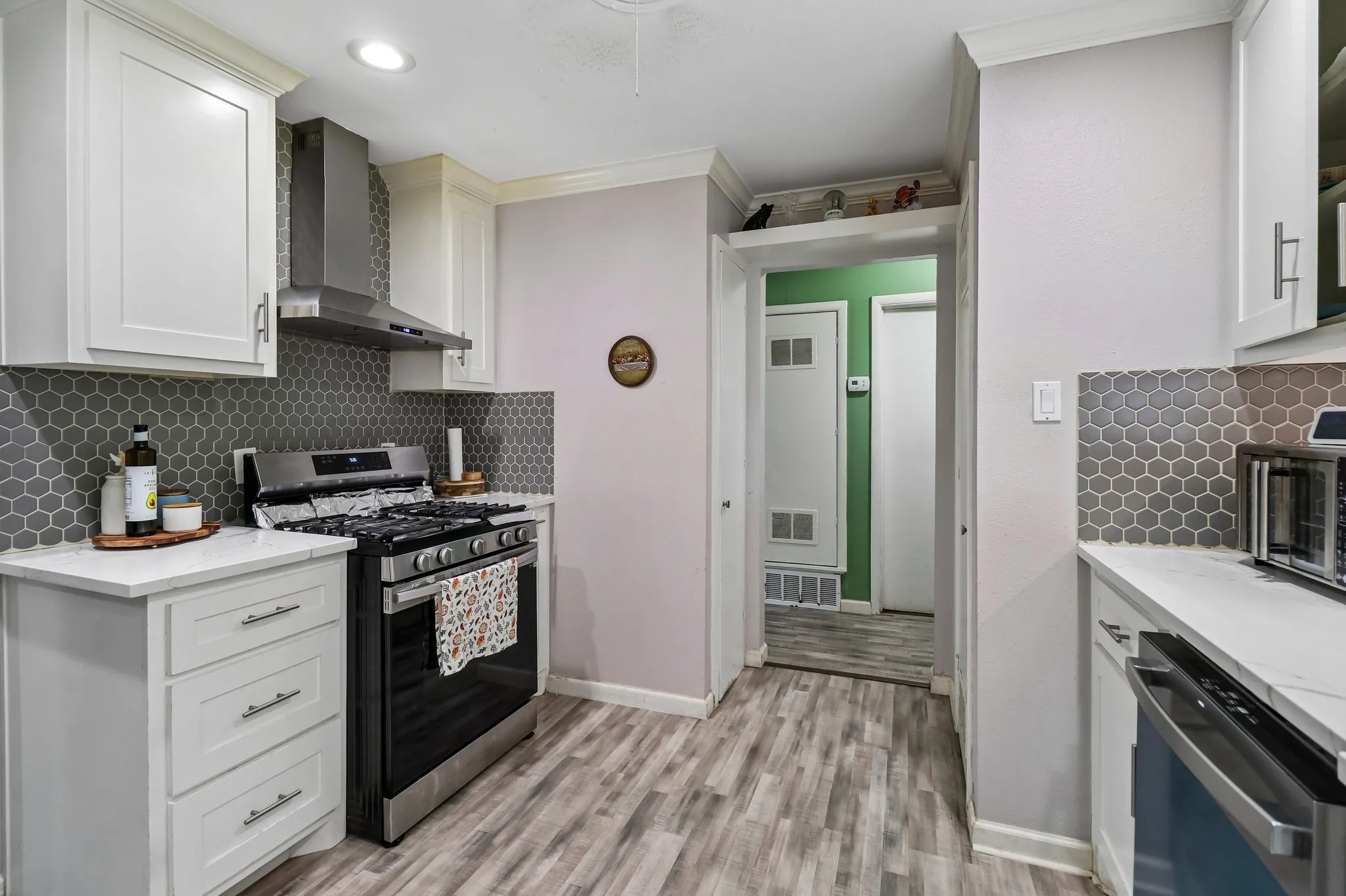 White shaker-style cabinetry, stainless steel range with matching hood, hexagonal tile backsplash, light-toned countertops, and wood-finish flooring