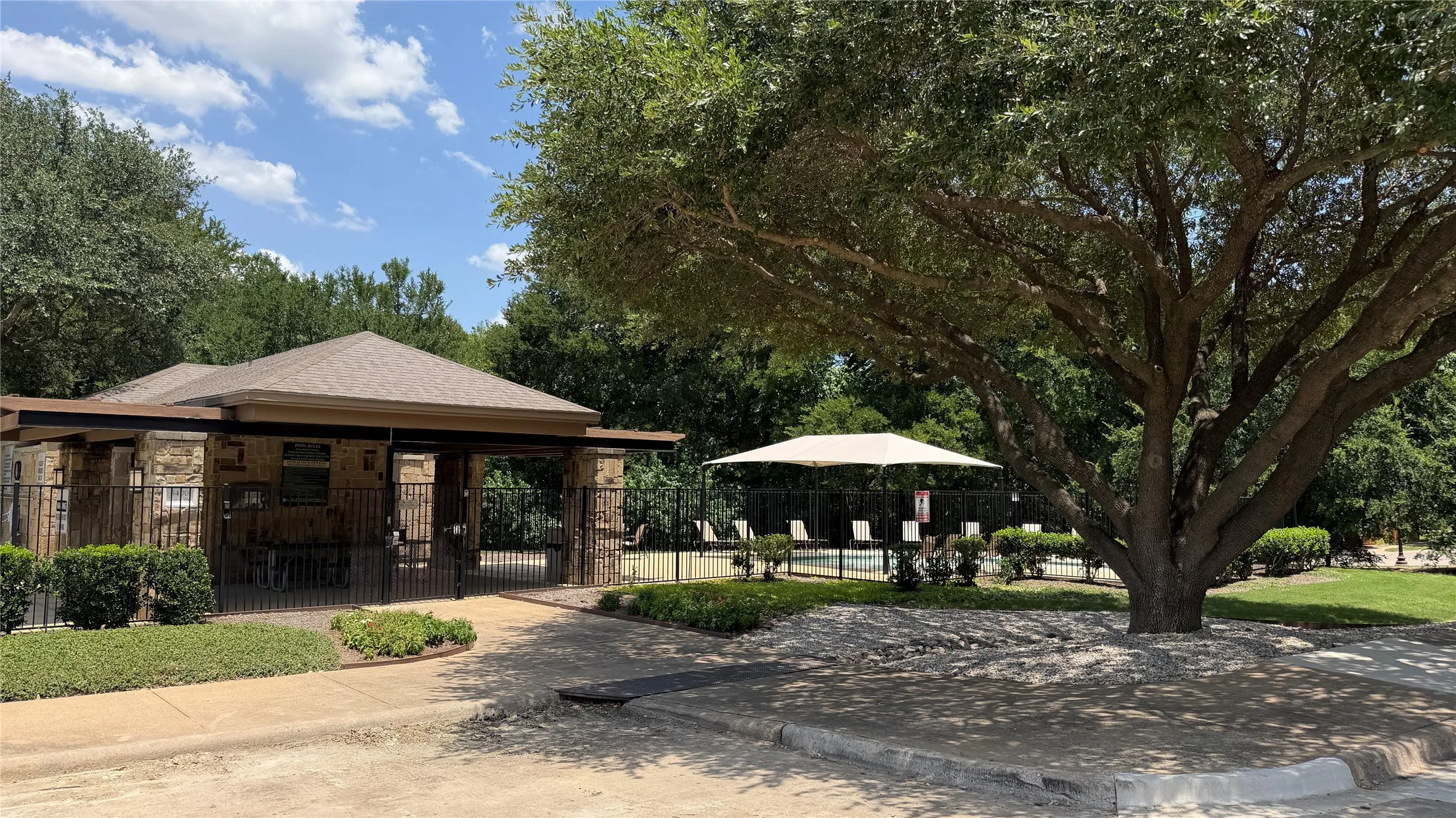 Community pool area featuring a stone and wrought-iron gated entry, mature landscaping, and an umbrella-shaded lounge area with poolside seating