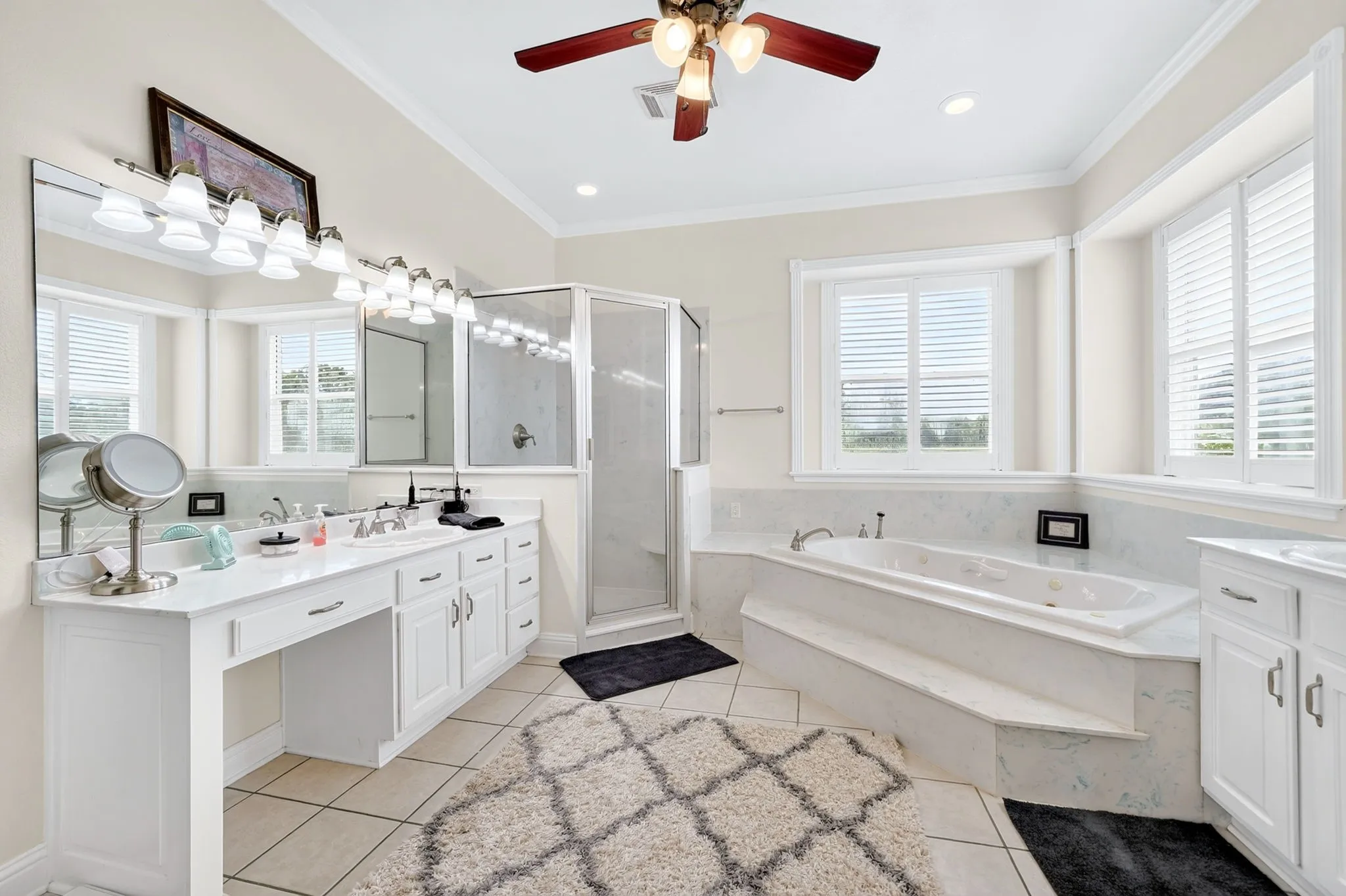 Bathroom featuring a built-in jetted tub with tile surround, a glass-enclosed shower, dual vanities with white cabinetry, and multiple windows with white louvered blinds