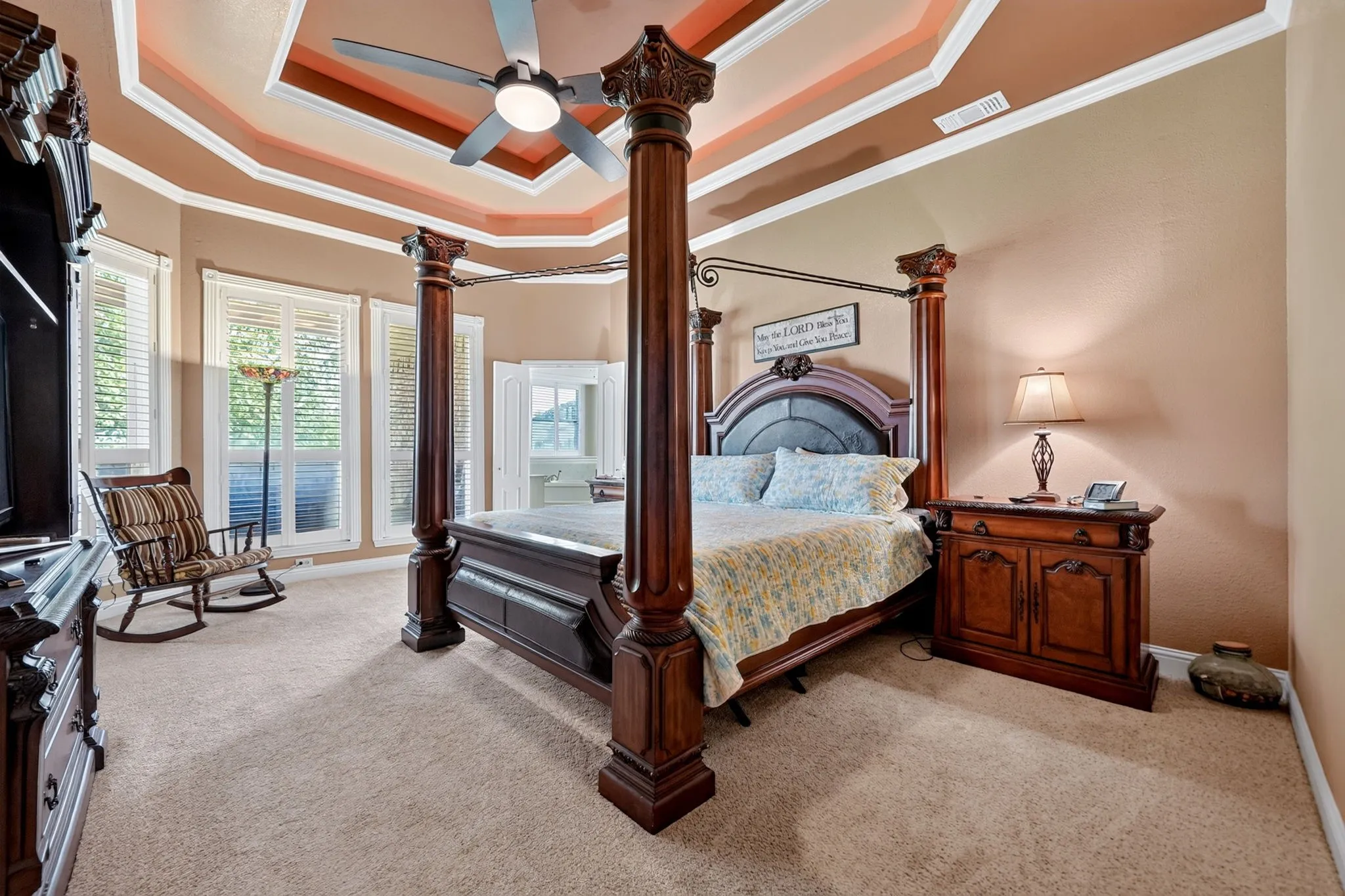 Spacious carpeted room featuring a multi-tiered tray ceiling with a ceiling fan, crown molding, and a bay window with plantation shutters