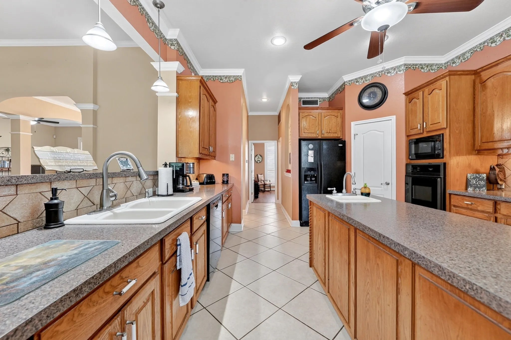 Expansive kitchen featuring a double basin sink, black appliances, a central island with an integrated sink, wood cabinetry, and tiled flooring