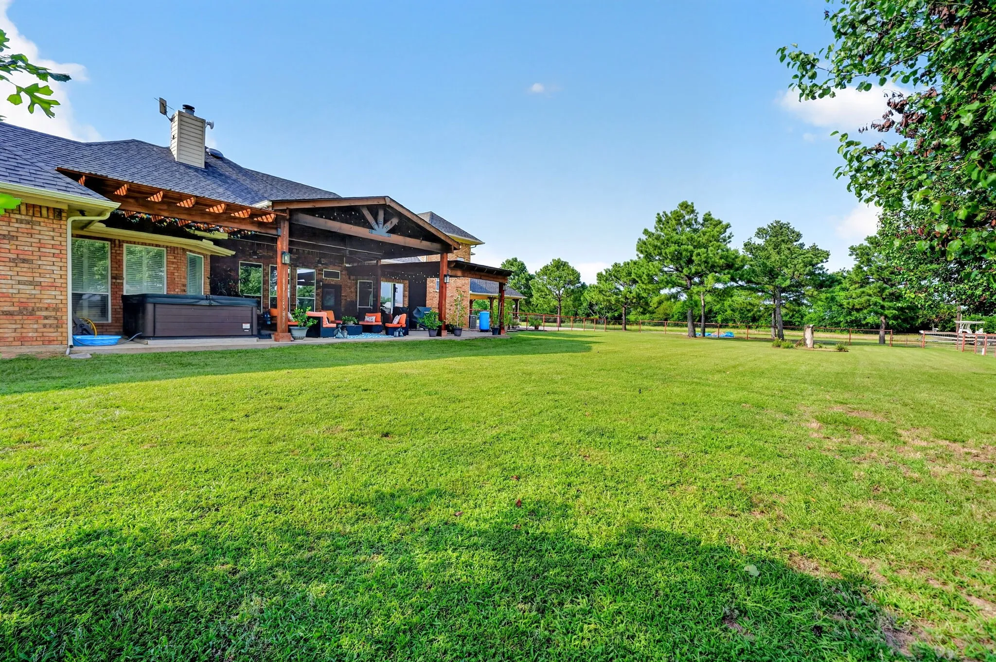 Spacious outdoor living area featuring a covered patio, extensive lawn, mature trees, and a brick exterior with a spa