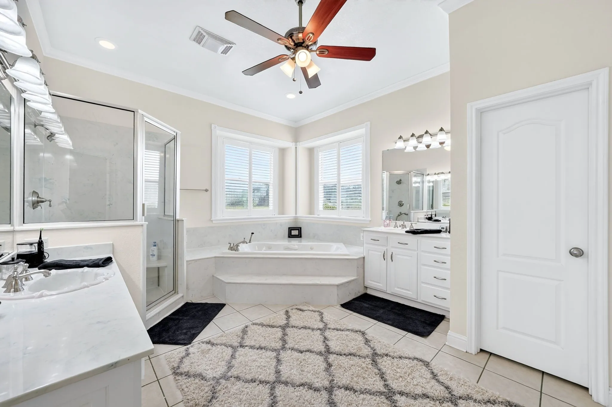 Spacious bathroom featuring a built-in jetted tub, dual vanity areas with white cabinetry, a glass-enclosed shower, and a ceiling fan with integrated lighting