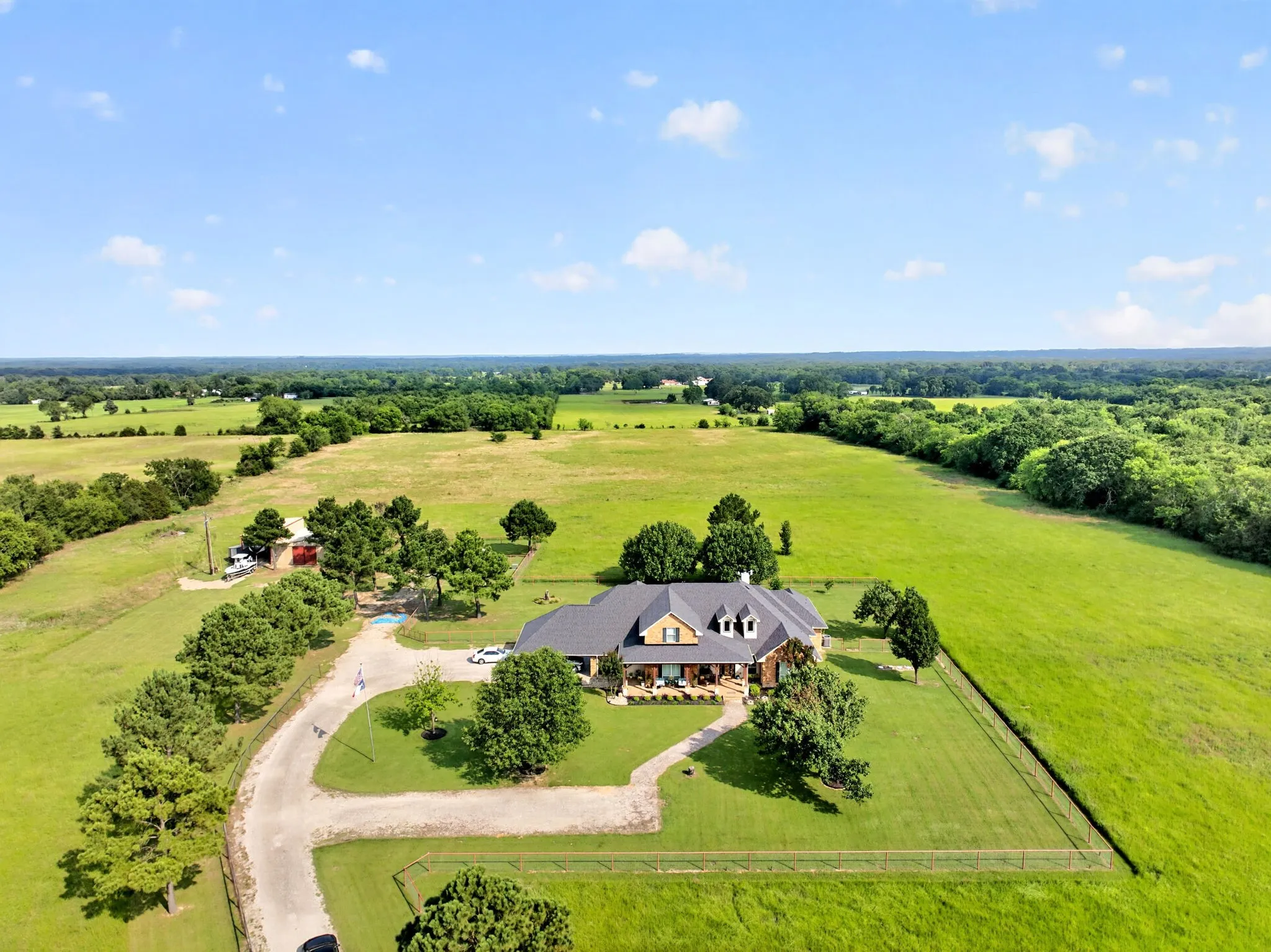 Expansive property featuring a stone and siding residence with a dark roof, surrounded by green fields and mature trees