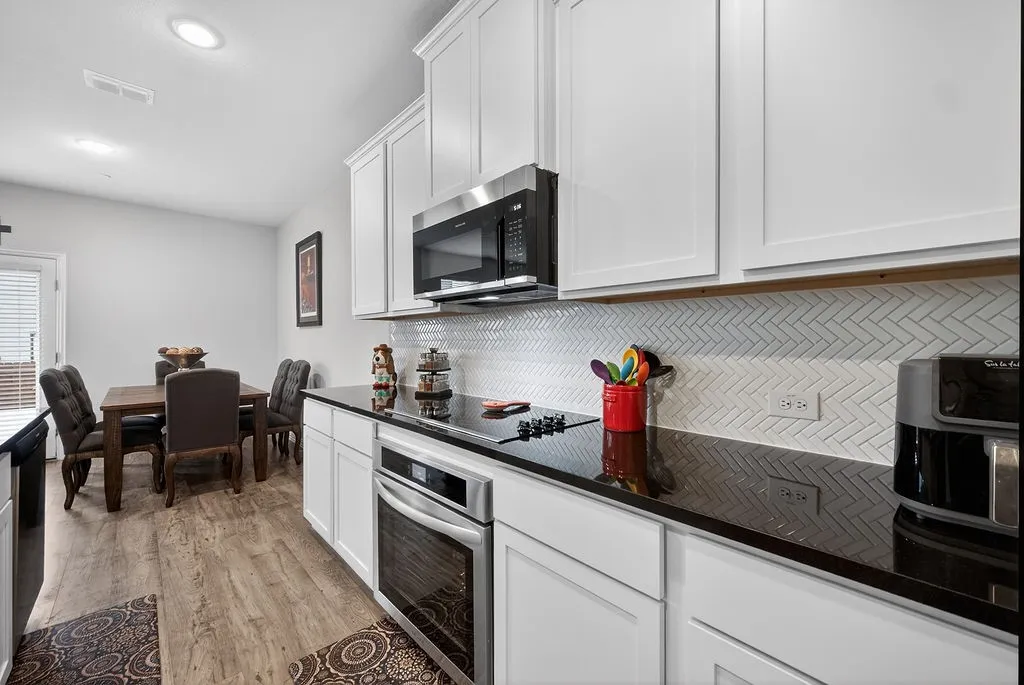 Kitchen featuring white shaker cabinetry, dark countertops, white herringbone backsplash, and stainless steel appliances including a built-in microwave and oven